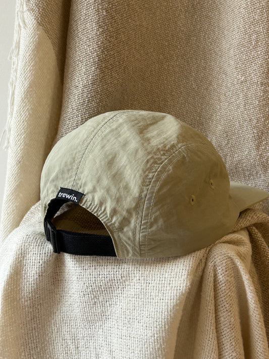 A beige 5-panel cap with a black adjustable strap and a small black brand tag reading 'brevin', styled on draped cream-colored textured fabric.