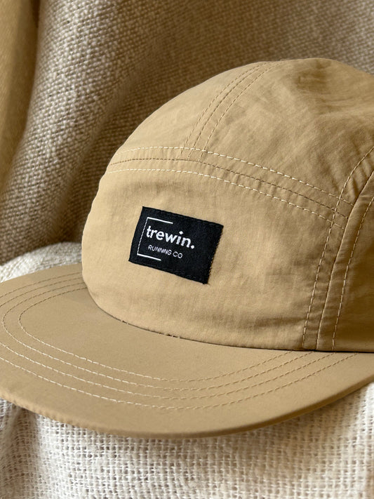 A close-up of a tan five-panel cap with a black rectangular patch on the front that reads 'trewin. RUNNING CO.