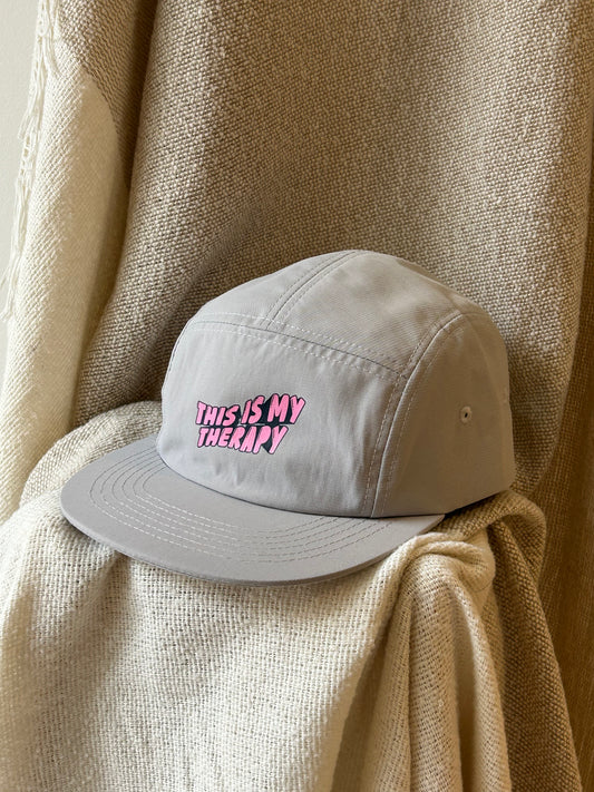 A light gray 5-panel cap with 'THIS IS MY THERAPY' embroidered in pink bubble letters on the front, resting on a textured beige blanket.