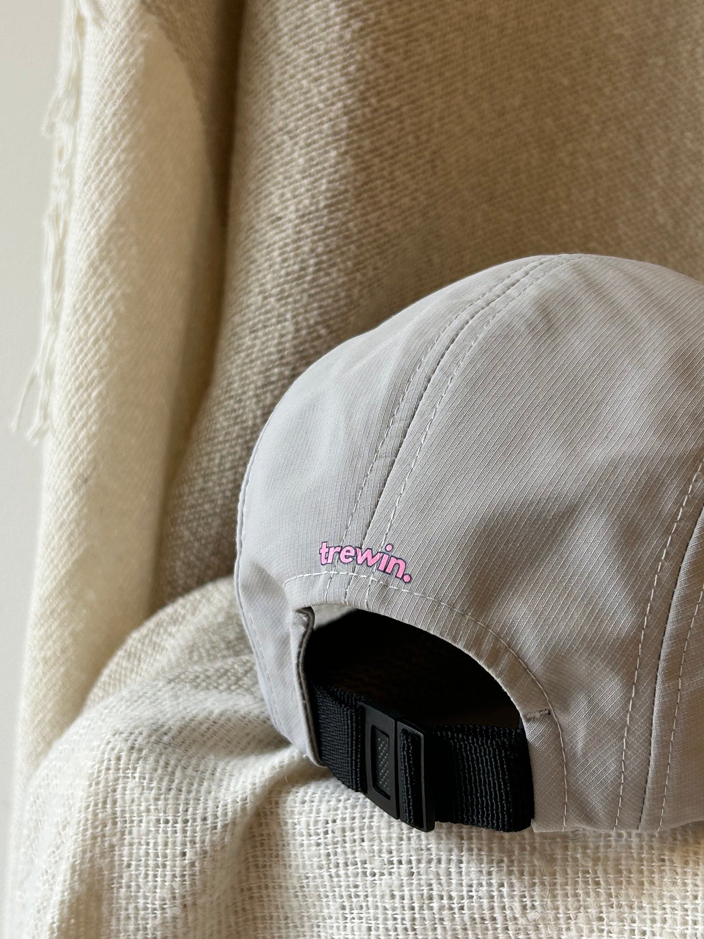 A close-up of the back of a light beige cap with a black adjustable strap and a small pink embroidered 'trewin.' logo, resting against a textured beige fabric backdrop.