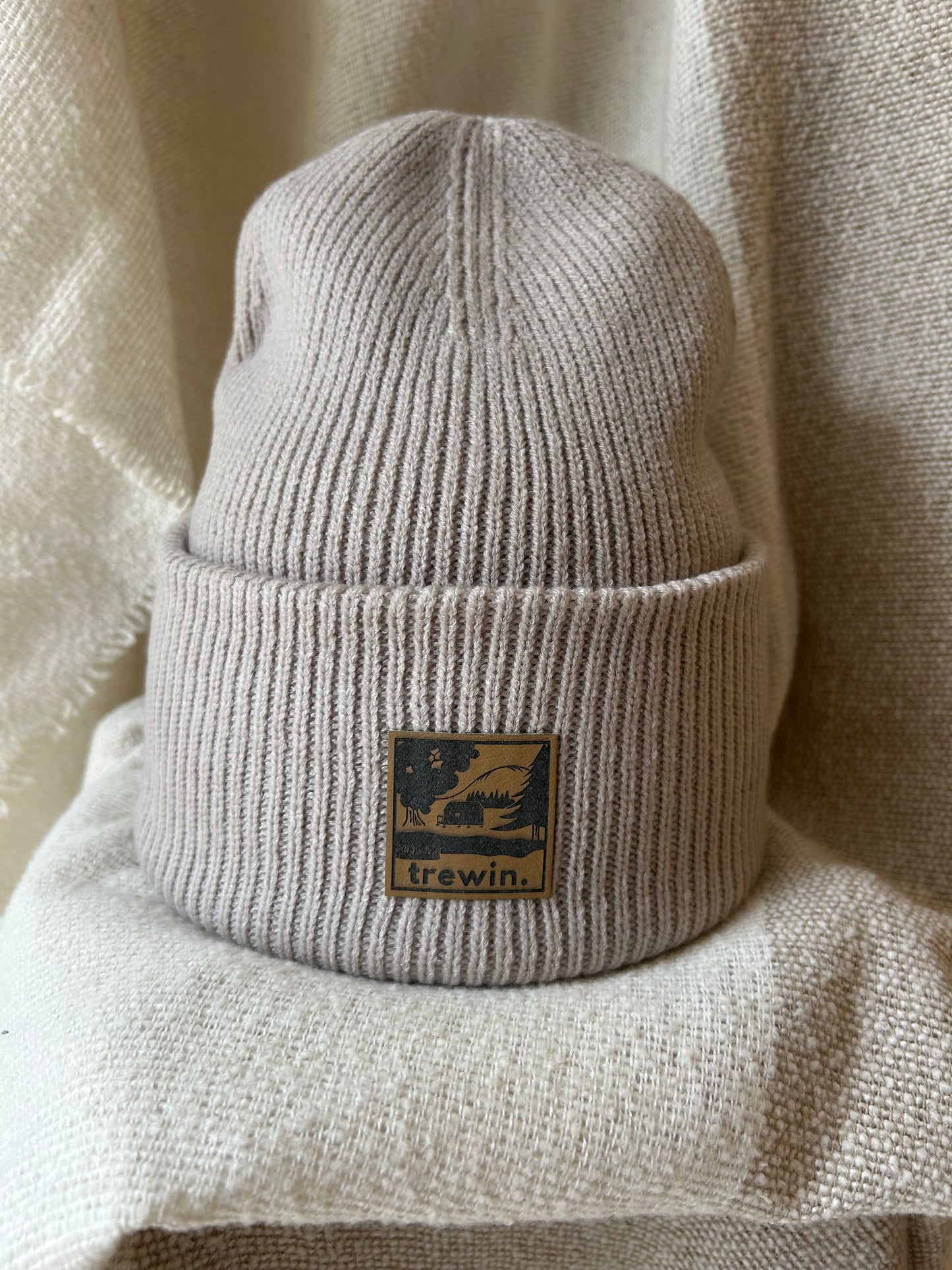 Close-up of a ribbed knit beanie in a warm taupe-beige color, featuring a square brown leather patch with a black graphic of a cabin in a forest and the brand name 'trewin.'.