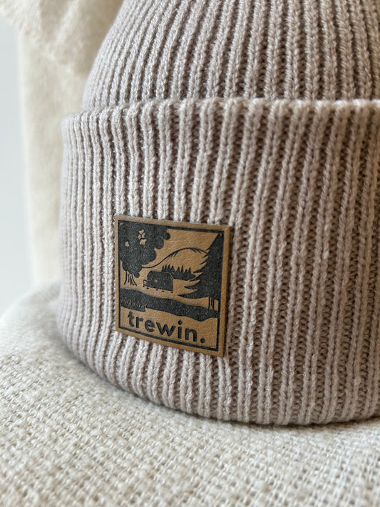 Close-up of a ribbed knit beanie in a warm taupe-beige color, featuring a square brown leather patch with a black graphic of a cabin in a forest and the brand name 'trewin.'.