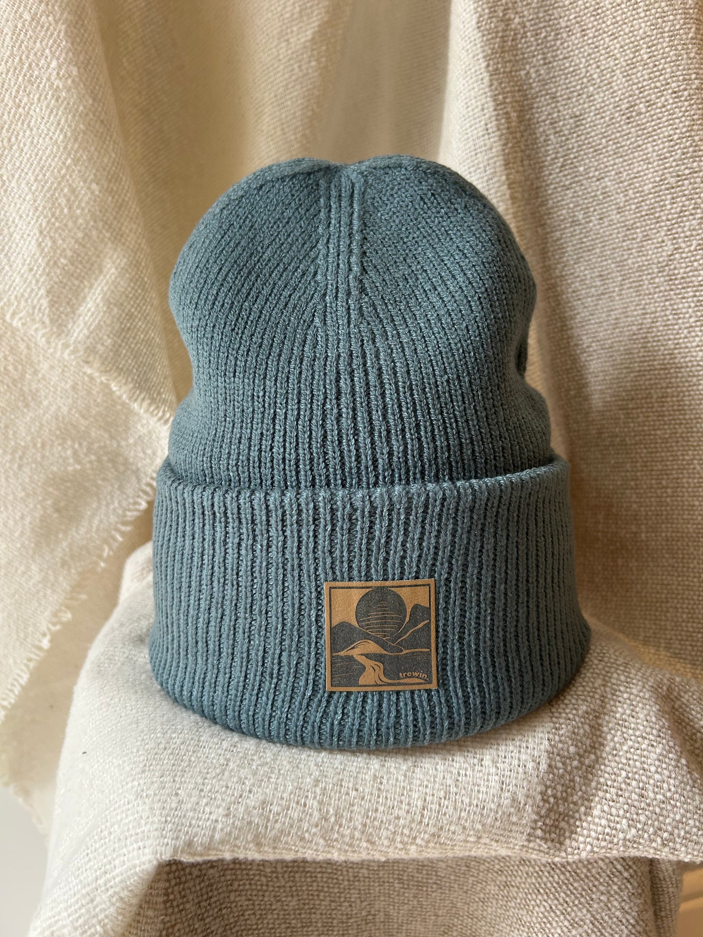 A dusty blue ribbed knit beanie hat with a tan square patch featuring a dark graphic logo, displayed on a textured beige fabric background.