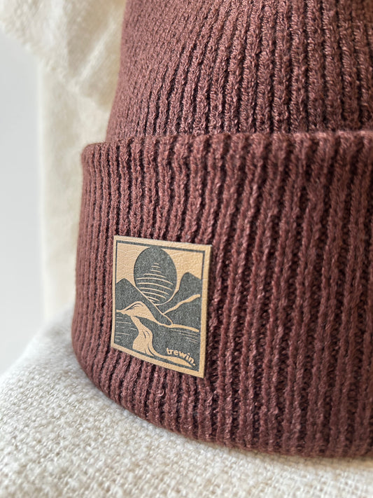 Close-up of a ribbed knit beanie in a dusty mauve-brown color, featuring a square tan patch with a dark graphic logo and the word 'yewwin'.