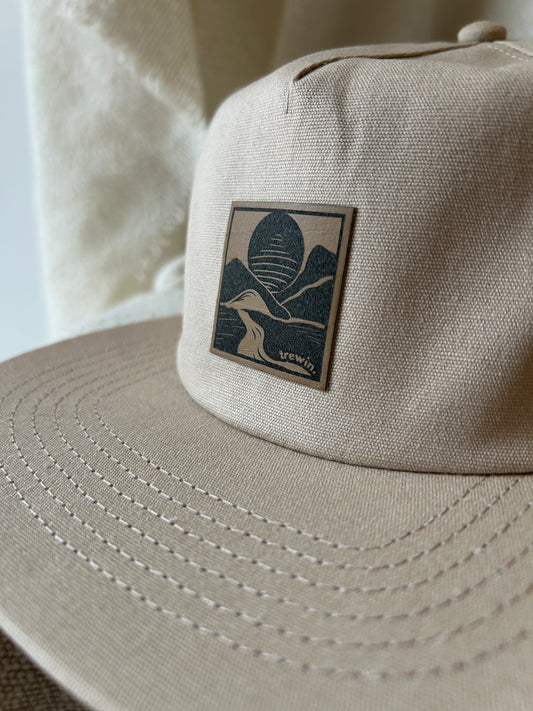 A close-up of a beige canvas trucker hat featuring a square patch on the front with a dark blue graphic of mountains and a bird, with the brand name 'U.S.