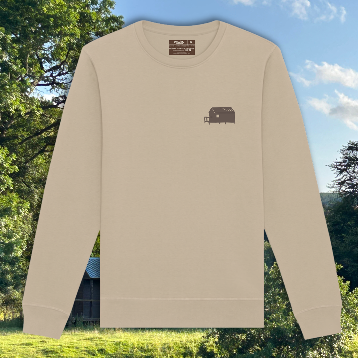 A beige long-sleeve crewneck sweatshirt with a small brown graphic of a small cabin on the left chest, displayed against a background of green trees and a blue sky.