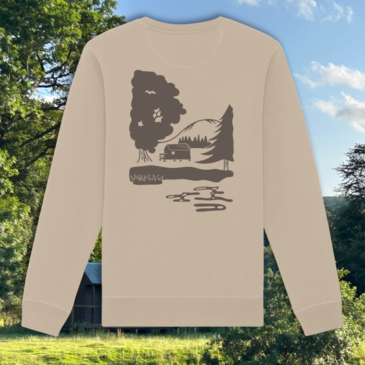 A beige long-sleeve crewneck sweatshirt with a dark brown graphic of a cabin, trees, and mountains, displayed against a background of a green outdoor landscape with trees and a blue sky.