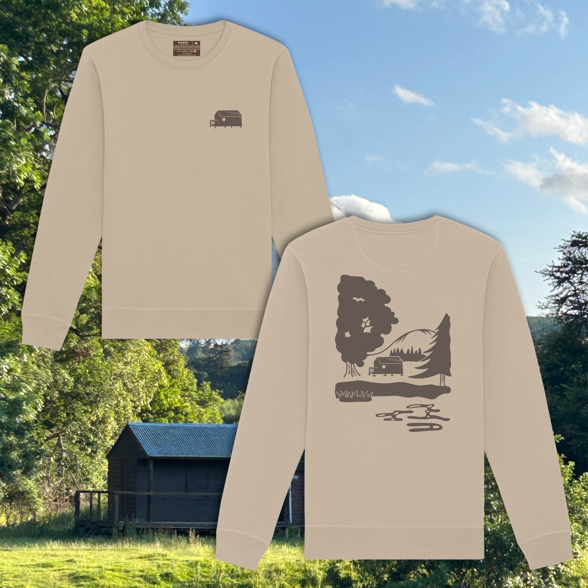 Two beige crewneck sweatshirts shown against a scenic outdoor background of trees, a cabin, and blue sky. The top sweatshirt features a small dark brown campsite graphic on the left chest, while the bottom sweatshirt displays a larger matching campsite graphic on the front.
