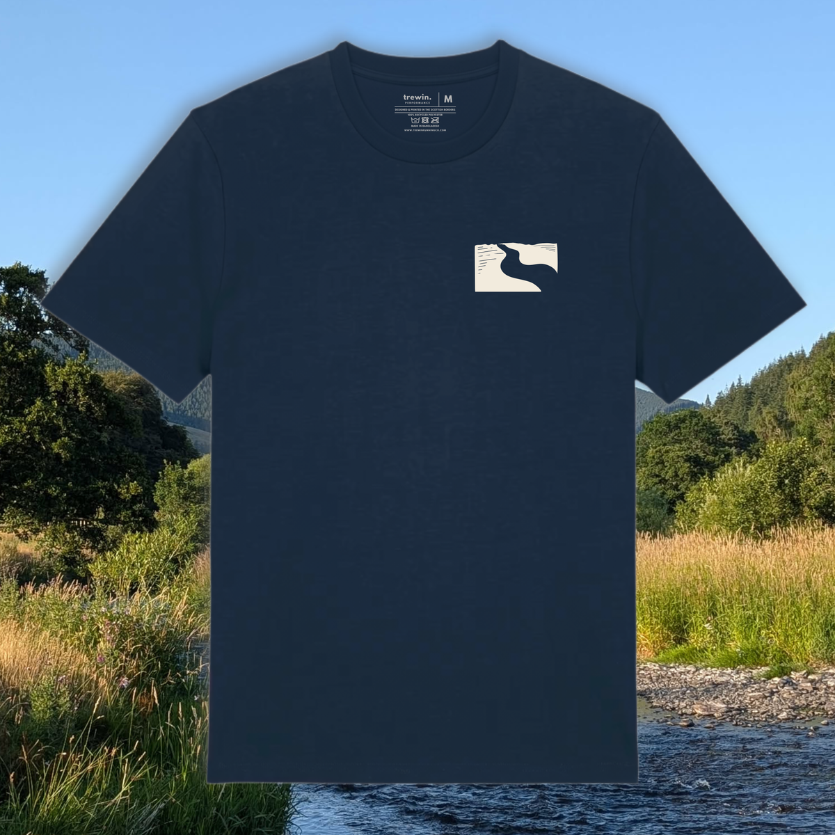 A dark navy blue crewneck t-shirt with a small white rectangular chest graphic, shown against a scenic outdoor background of a river and grassy landscape.