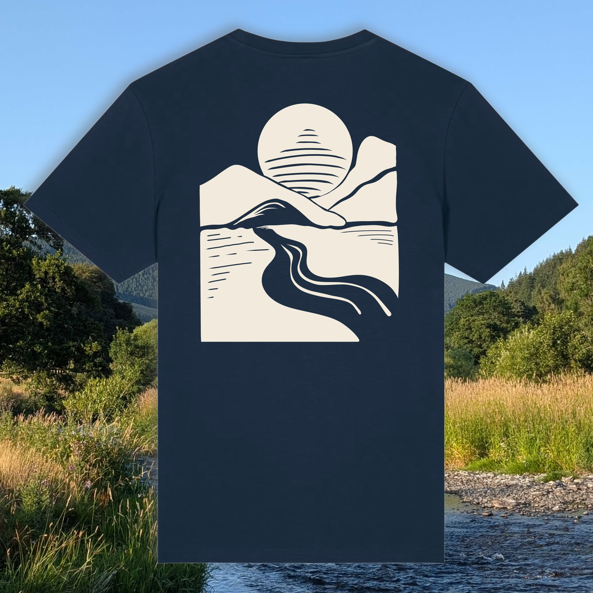 A navy blue short-sleeve t-shirt with a cream-colored minimalist graphic of a mountain, river, and sun on the back, displayed against a scenic outdoor background of a river and grassy fields.