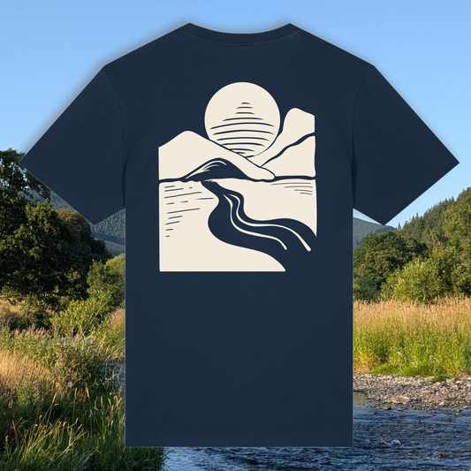 A navy blue short-sleeve t-shirt with a cream-colored minimalist graphic of a mountain, river, and sun on the back, displayed against a scenic outdoor background of a river and grassy fields.