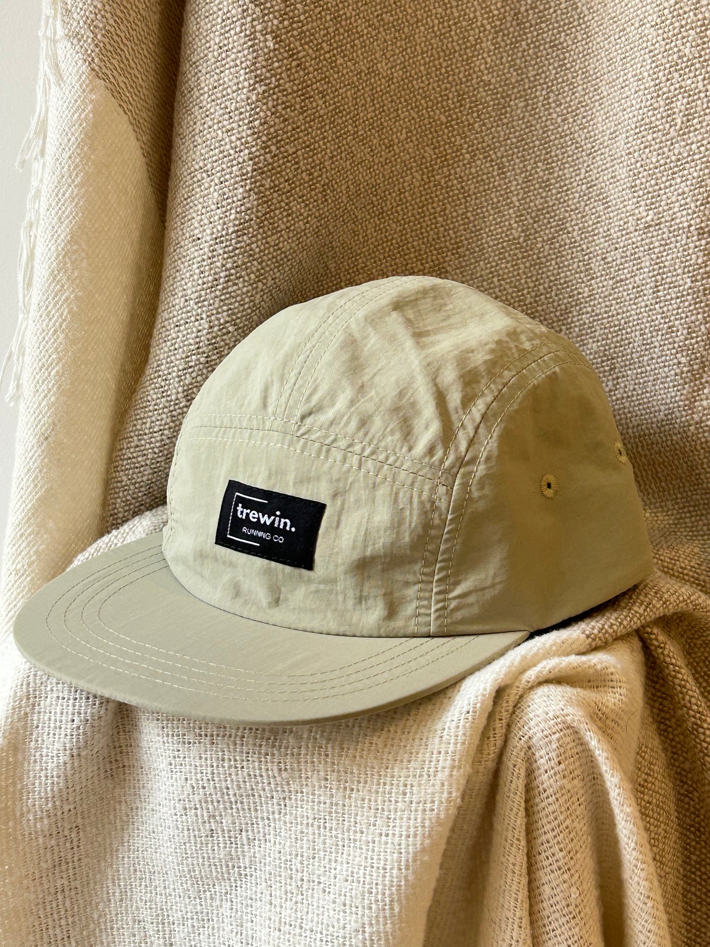 A light beige 5-panel camp cap with a black rectangular patch on the front reading 'trewin. LOS ANGELES CA', displayed on draped beige fabric.
