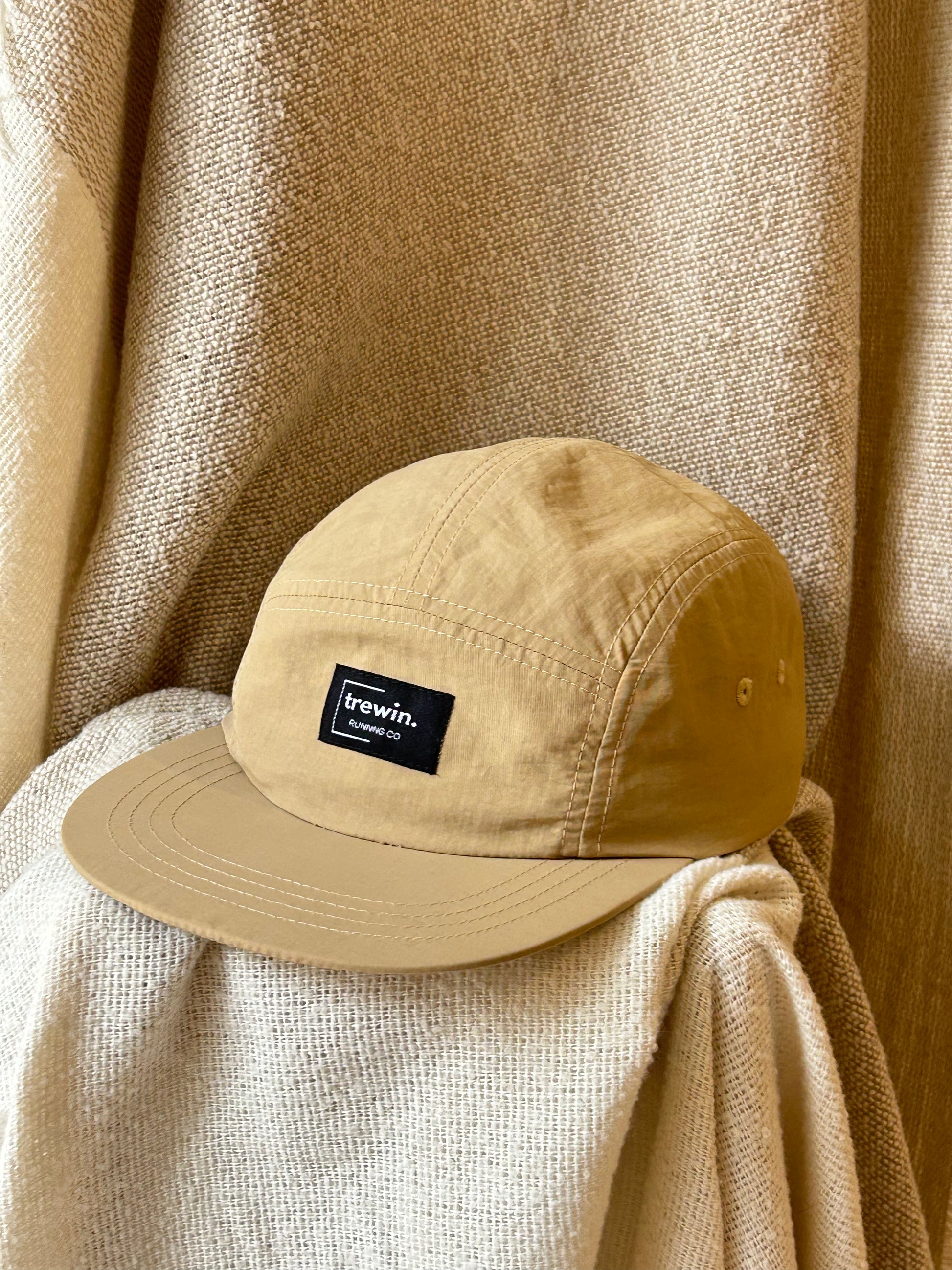 A tan 5-panel cap with a black 'trewin.' brand patch on the front, resting on draped beige fabric against a matching textured backdrop.