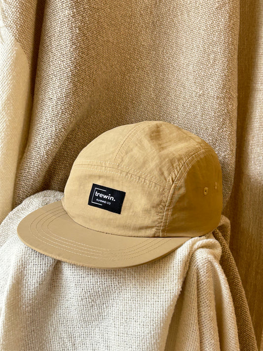 A tan 5-panel cap with a black 'trewin.' brand patch on the front, resting on draped beige fabric against a matching textured backdrop.