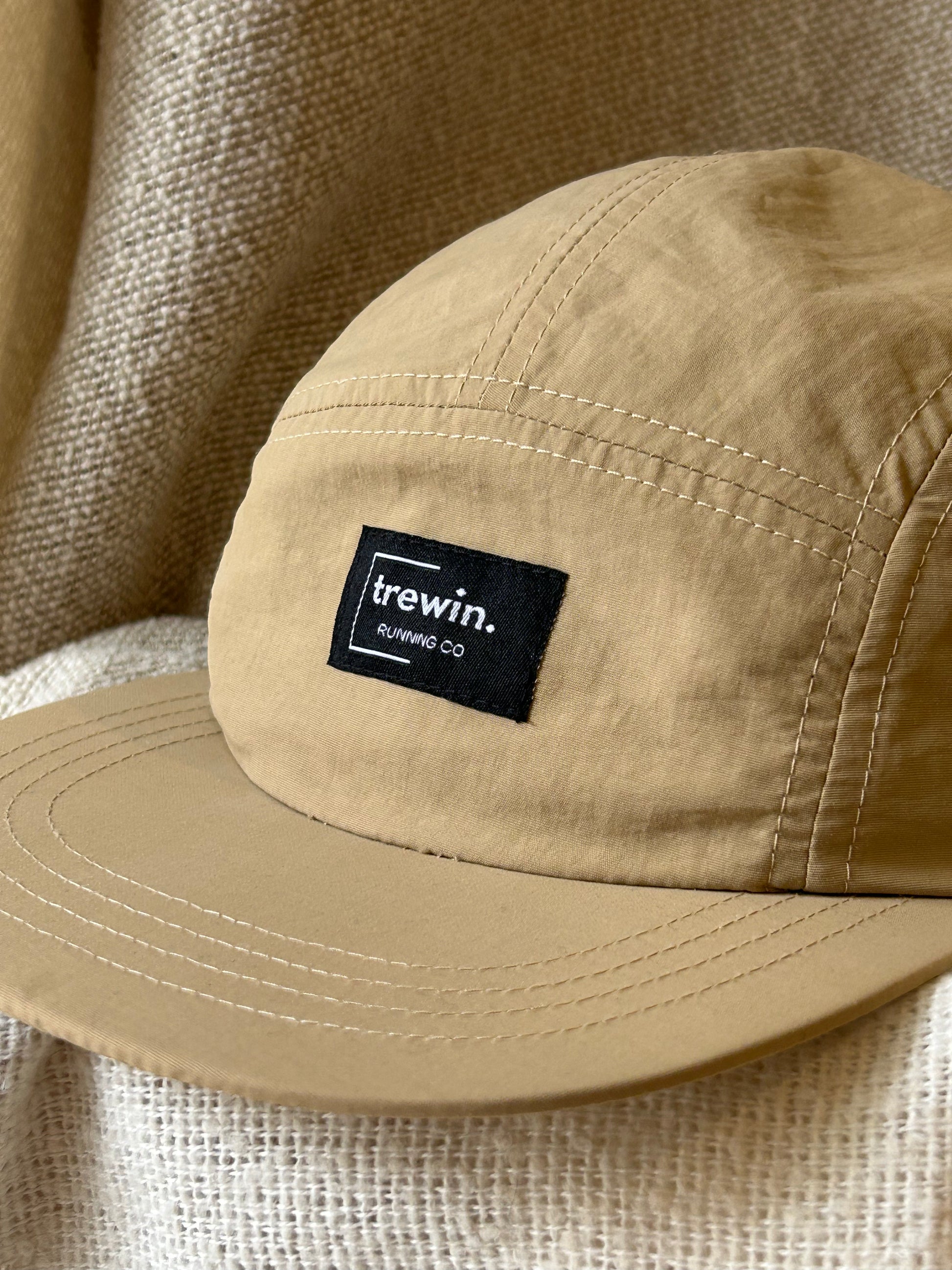 A close-up of a tan five-panel cap with a black rectangular patch on the front that reads 'trewin. RUNNING CO.