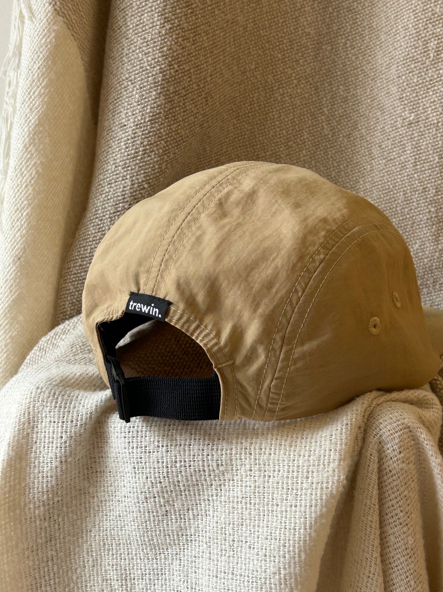 A tan cotton cap with a black adjustable strap and a small black 'trewin' brand tag, resting on a textured cream-colored fabric backdrop.