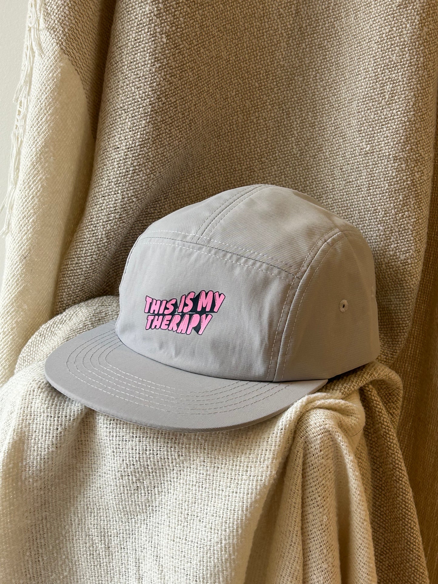 A light gray 5-panel cap with 'THIS IS MY THERAPY' embroidered in pink bubble letters on the front, resting on a textured beige blanket.