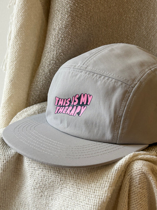 A light gray five-panel cap with the phrase 'THIS IS MY THERAPY' embroidered in pink bubble letters on the front, resting on a textured beige knit blanket.