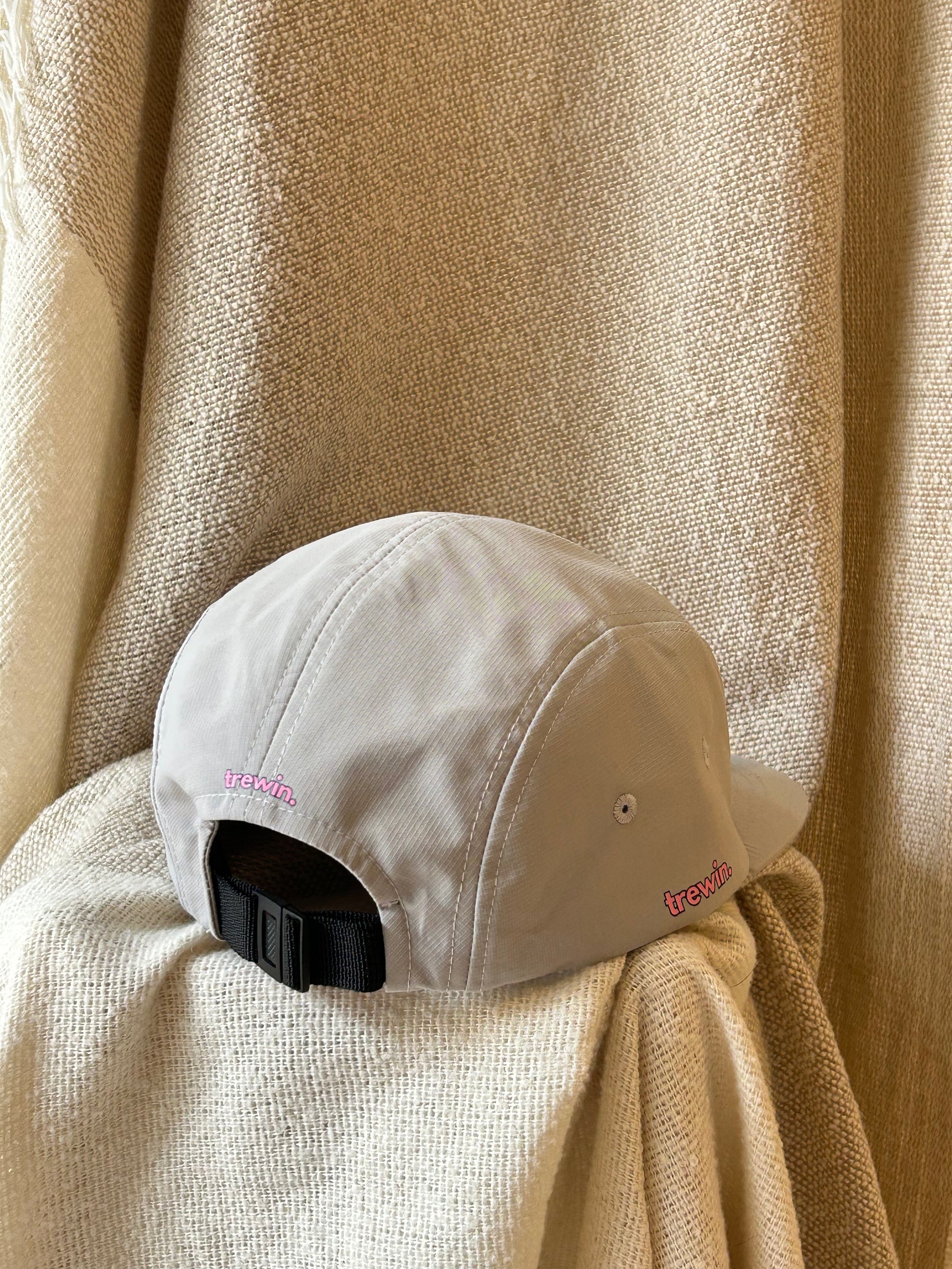 A light beige 5-panel baseball cap with a black adjustable strap and pink embroidered 'trewin' logos on the side, resting on a textured beige fabric backdrop.