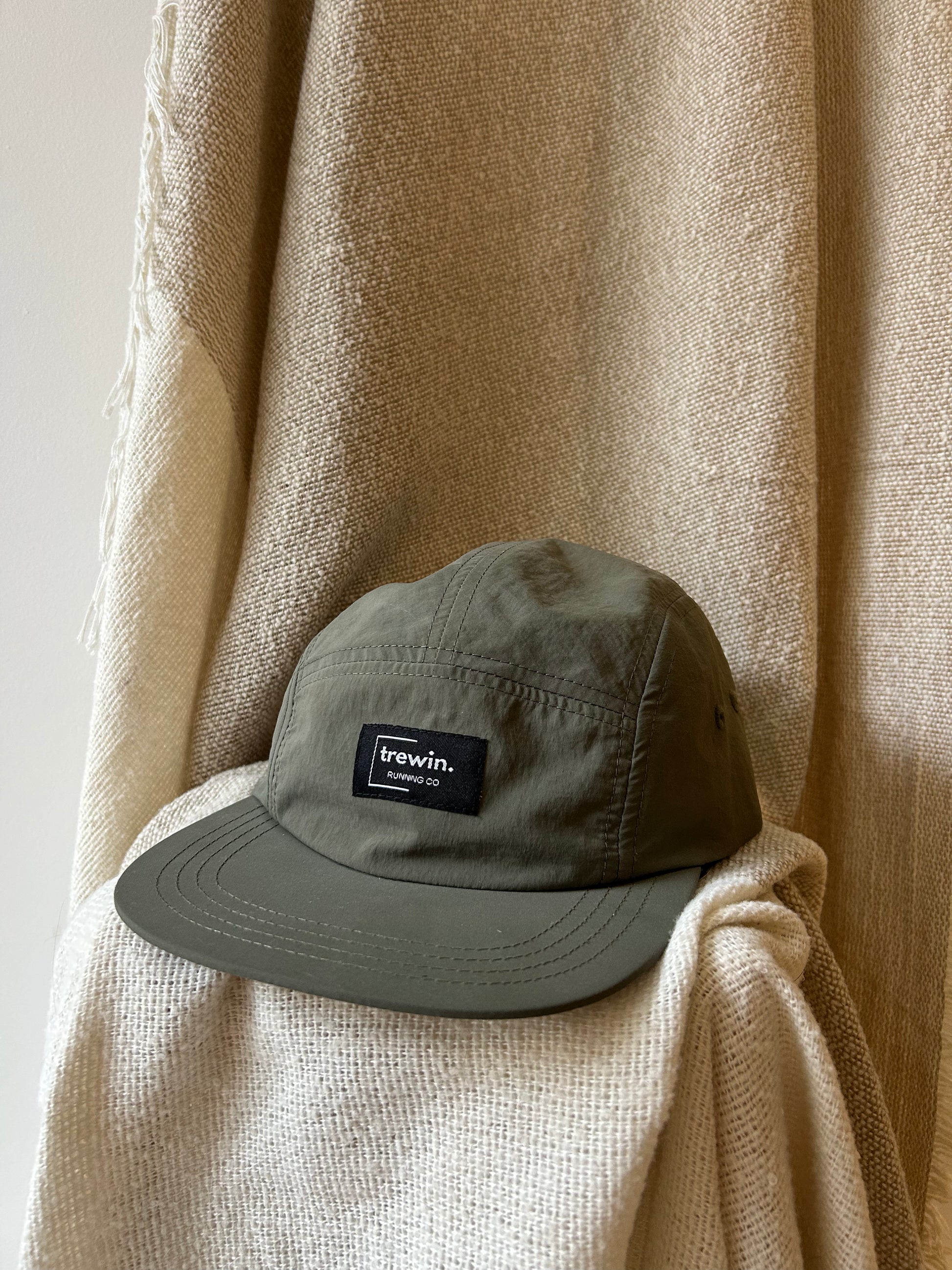 An olive green 5-panel cap with a black rectangular 'trewin. London Co.
