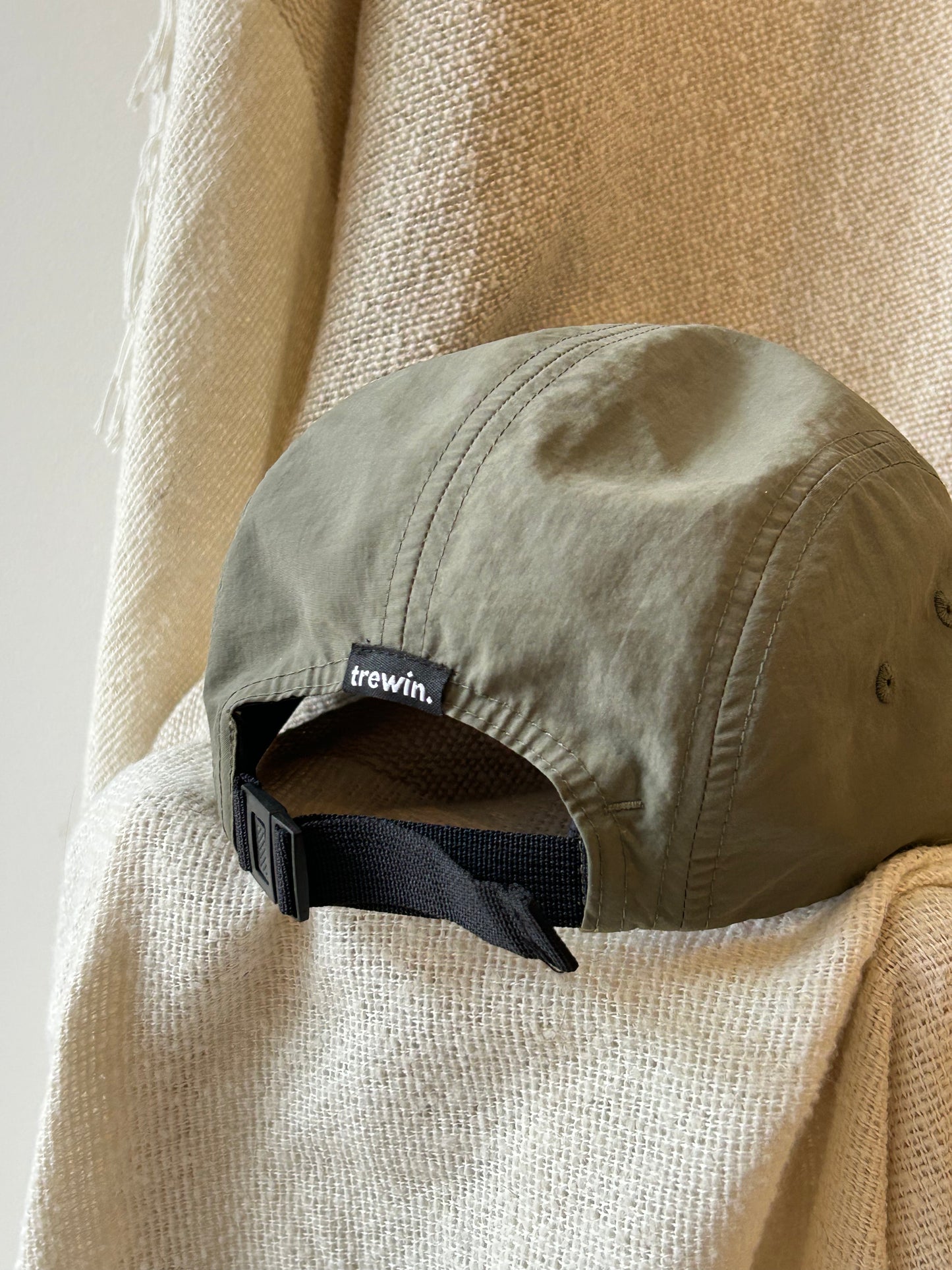 An olive green 5-panel cap with a black adjustable strap and a small black 'trewin.' brand tag, resting on a textured beige woven blanket.