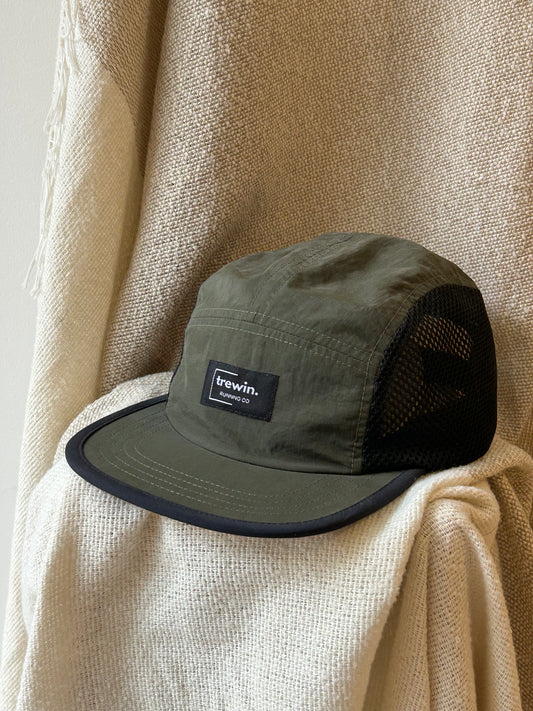 An olive green 5-panel trucker cap with black mesh back panels and a black visor trim, featuring a 'frewin. London' brand patch on the front, styled on a textured cream woven fabric backdrop.