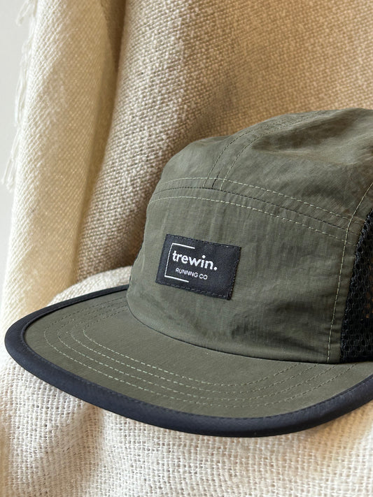 An olive green Trewin 5-panel camp cap with a black mesh back and black visor trim, resting on a textured beige woven blanket. The front features a black patch with the white 'trewin RUNNING CO' logo.