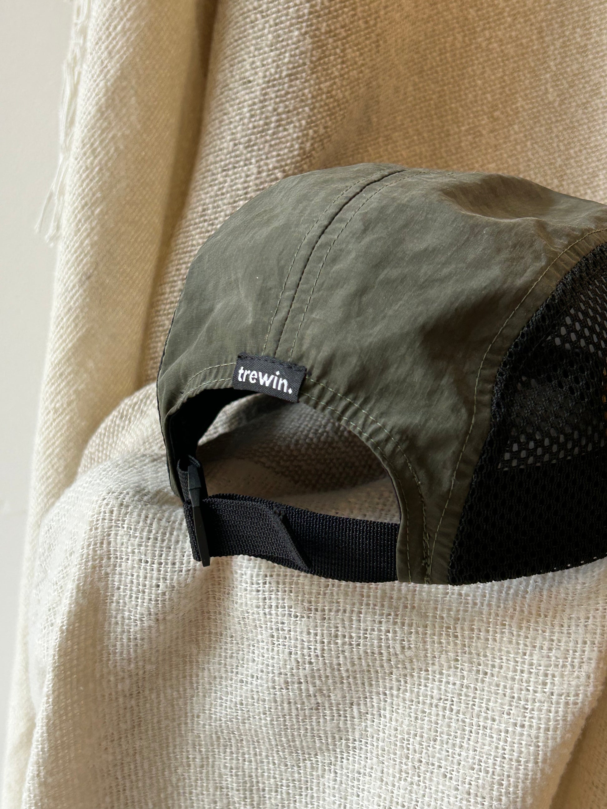 An olive green trucker-style cap with a black mesh back and an adjustable black strap, resting on a textured beige fabric. The brand name 'trewin.