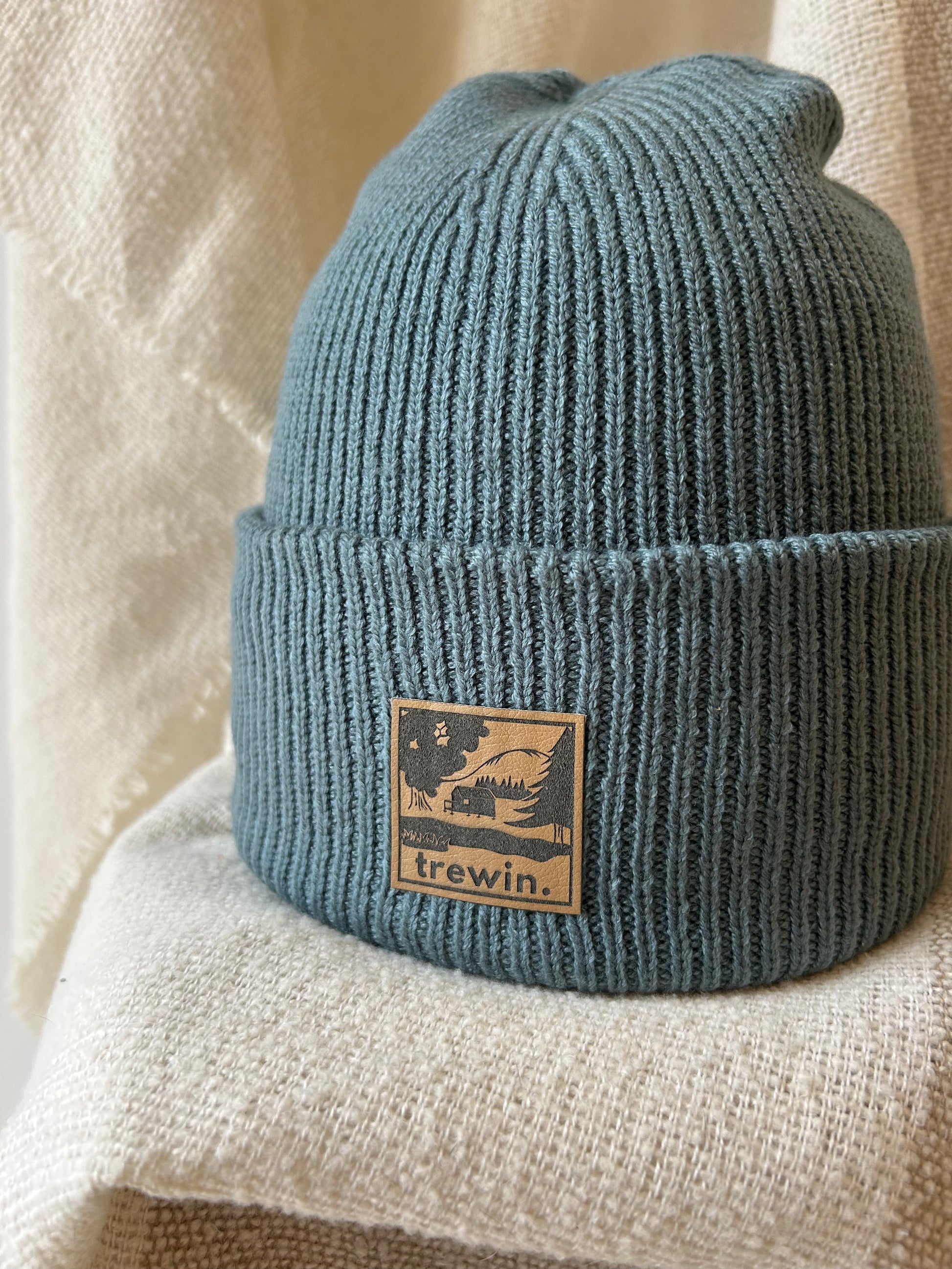 A ribbed knit beanie hat in a muted dusty blue-green color, featuring a tan square patch on the front with a graphic and the brand name 'trewin.' It is styled on a textured cream fabric surface.