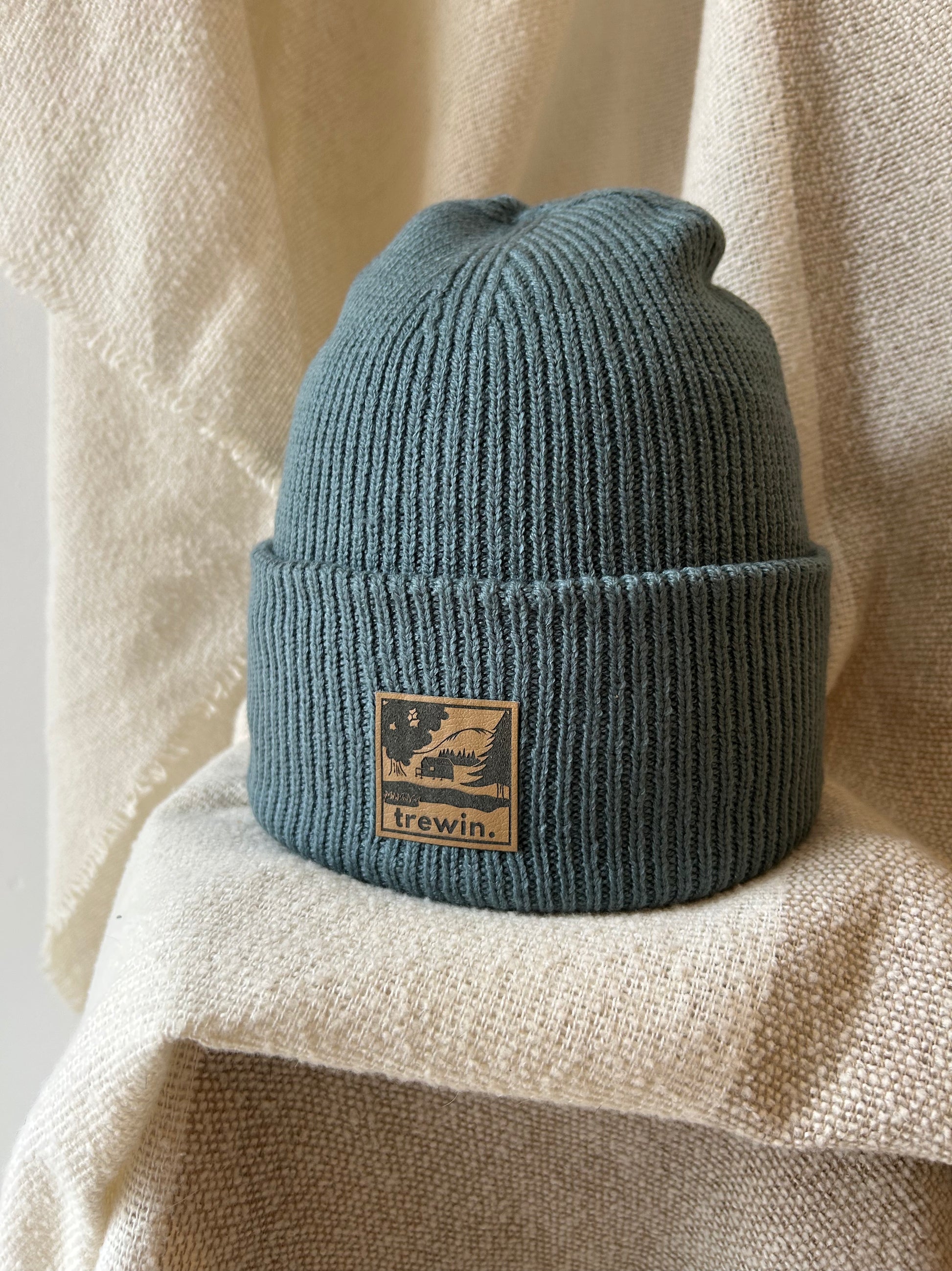 A ribbed knit beanie hat in a muted blue-green color, featuring a tan rectangular patch on the front cuff with the brand name 'trewin.' and a graphic of a dog on a leash.