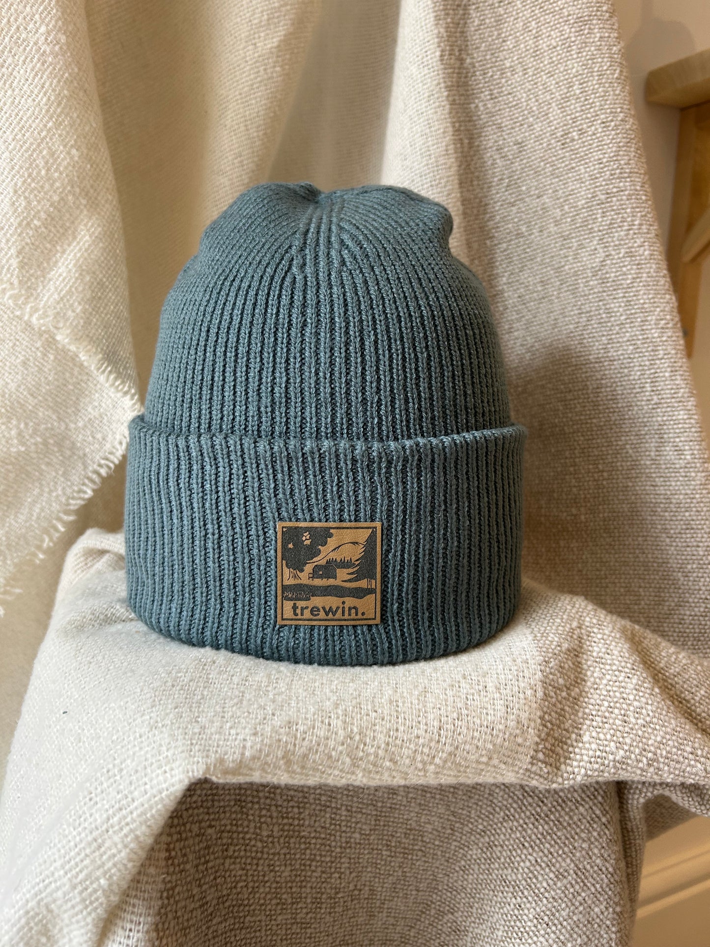 A dusty blue ribbed knit beanie hat with a tan square patch on the front that reads 'trewin.' and shows a small mountain graphic, displayed on draped beige fabric.