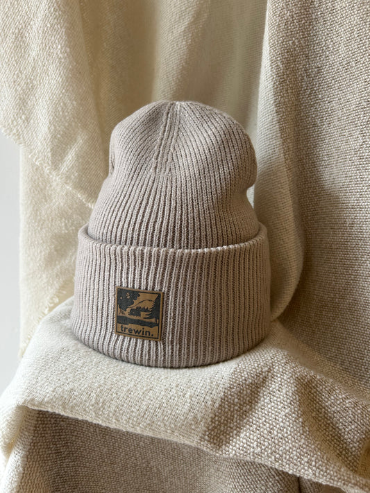 A ribbed knit beanie hat in a light taupe-beige color, featuring a square brown leather patch on the front cuff with the brand name 'KAVUIN' and a mountain graphic. The hat is styled on a draped, textured beige fabric background.