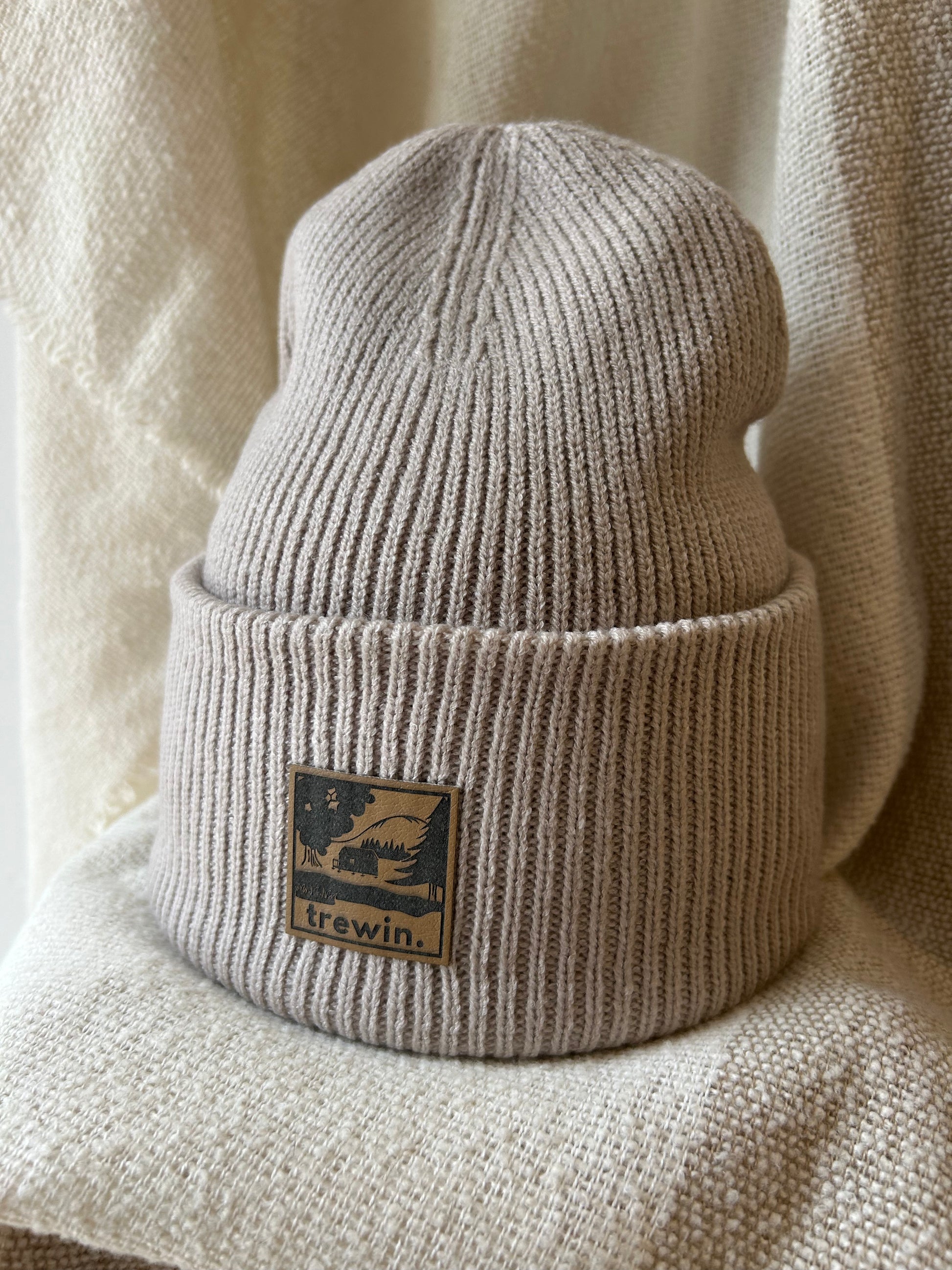 Close-up of a ribbed knit beanie in a warm taupe-beige color, featuring a square brown leather patch with a black graphic of a cabin in a forest and the brand name 'trewin.'.