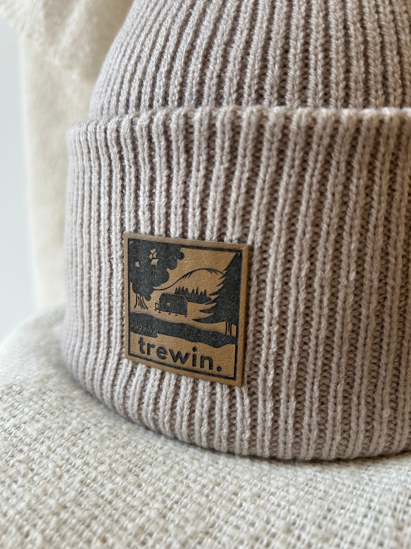 Close-up of a ribbed knit beanie in a warm taupe-beige color, featuring a square brown leather patch with a black graphic of a cabin in a forest and the brand name 'trewin.'.