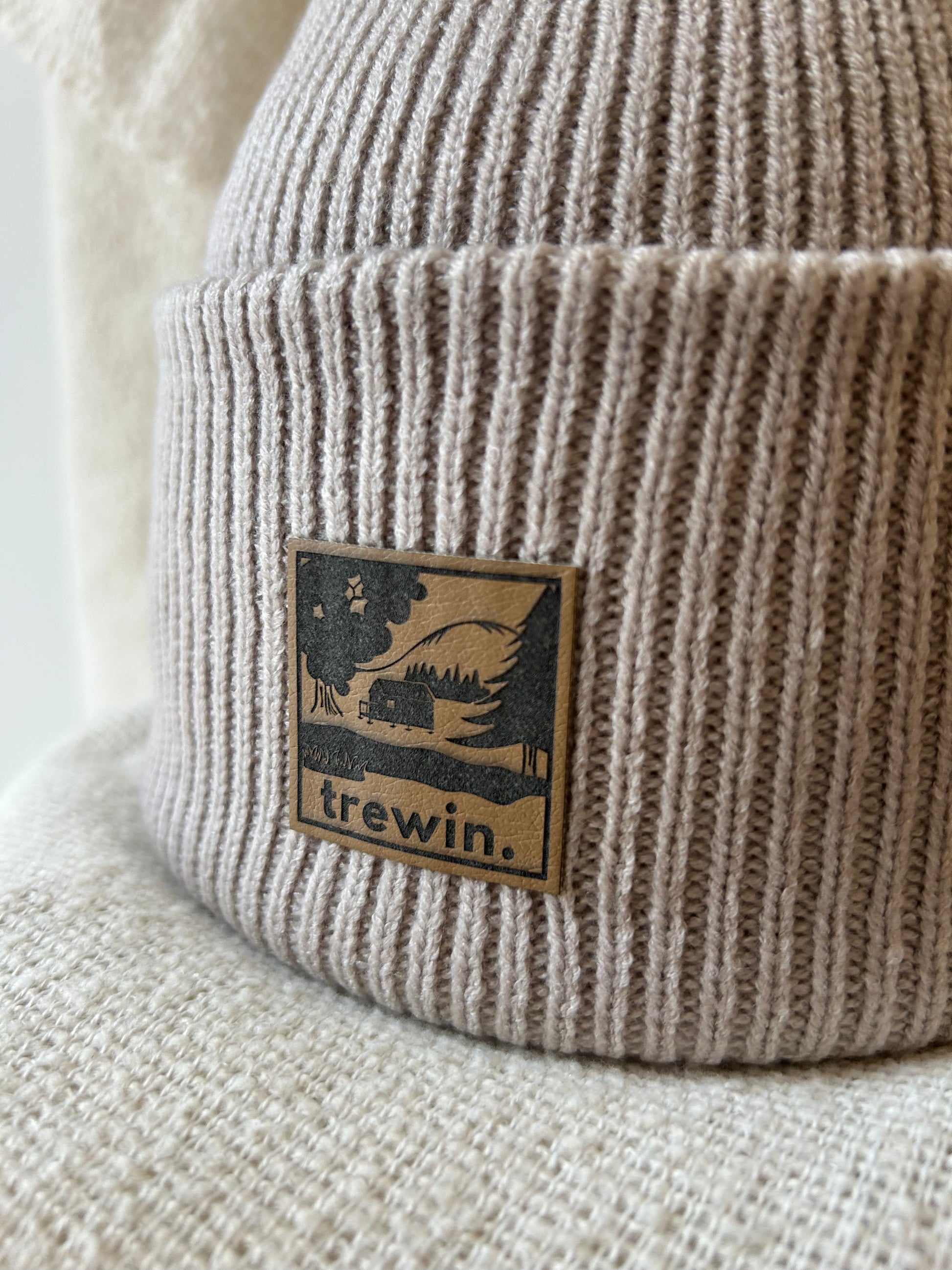 Close-up of a ribbed knit beanie in a warm taupe-beige color, featuring a square brown leather patch with a black graphic of a cabin in a forest and the brand name 'trewin.'.