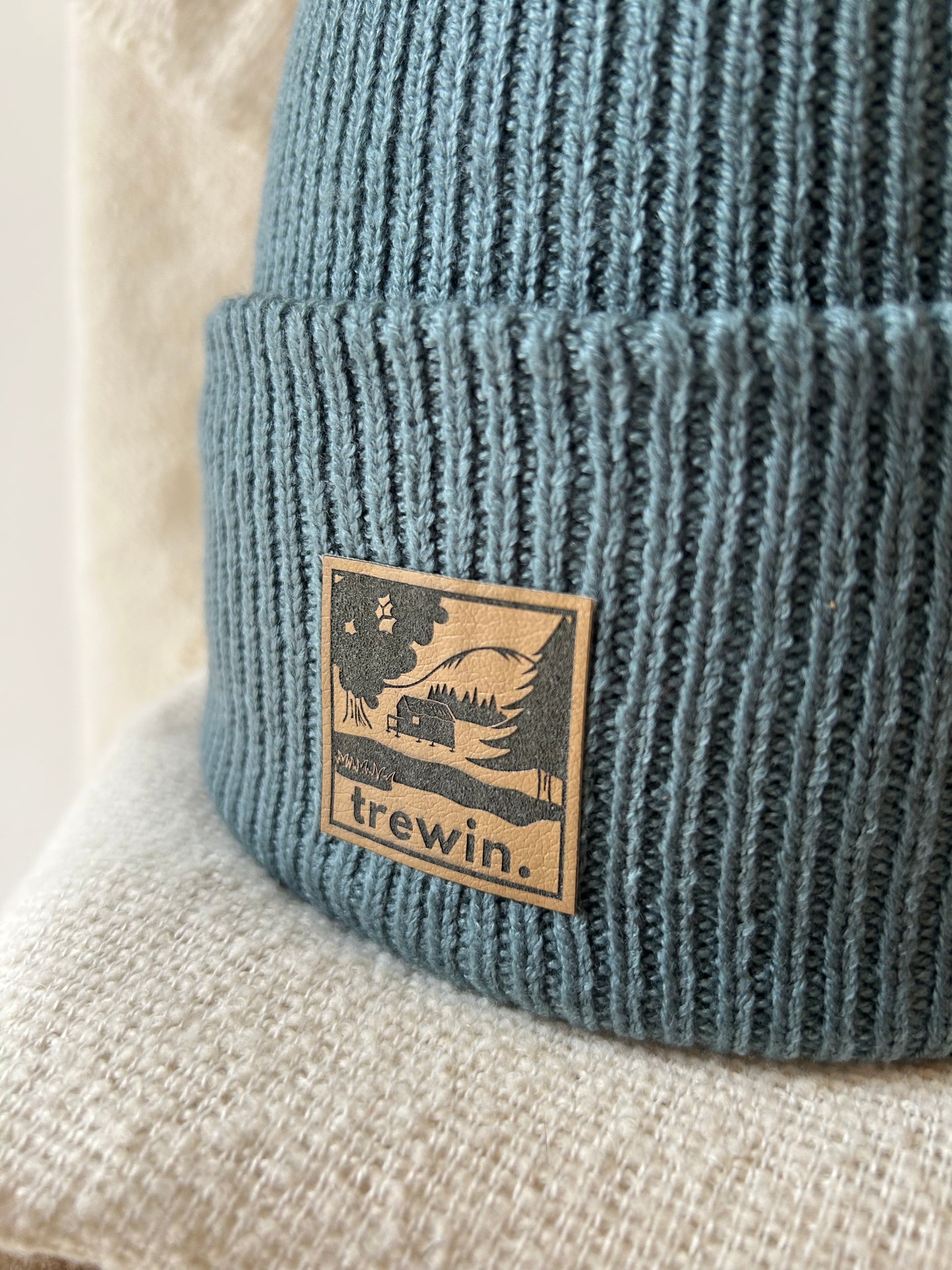 A close-up of a ribbed knit beanie in muted blue-gray, featuring a square tan leather patch on the cuff with a black forest illustration and the word 'trewin.'.