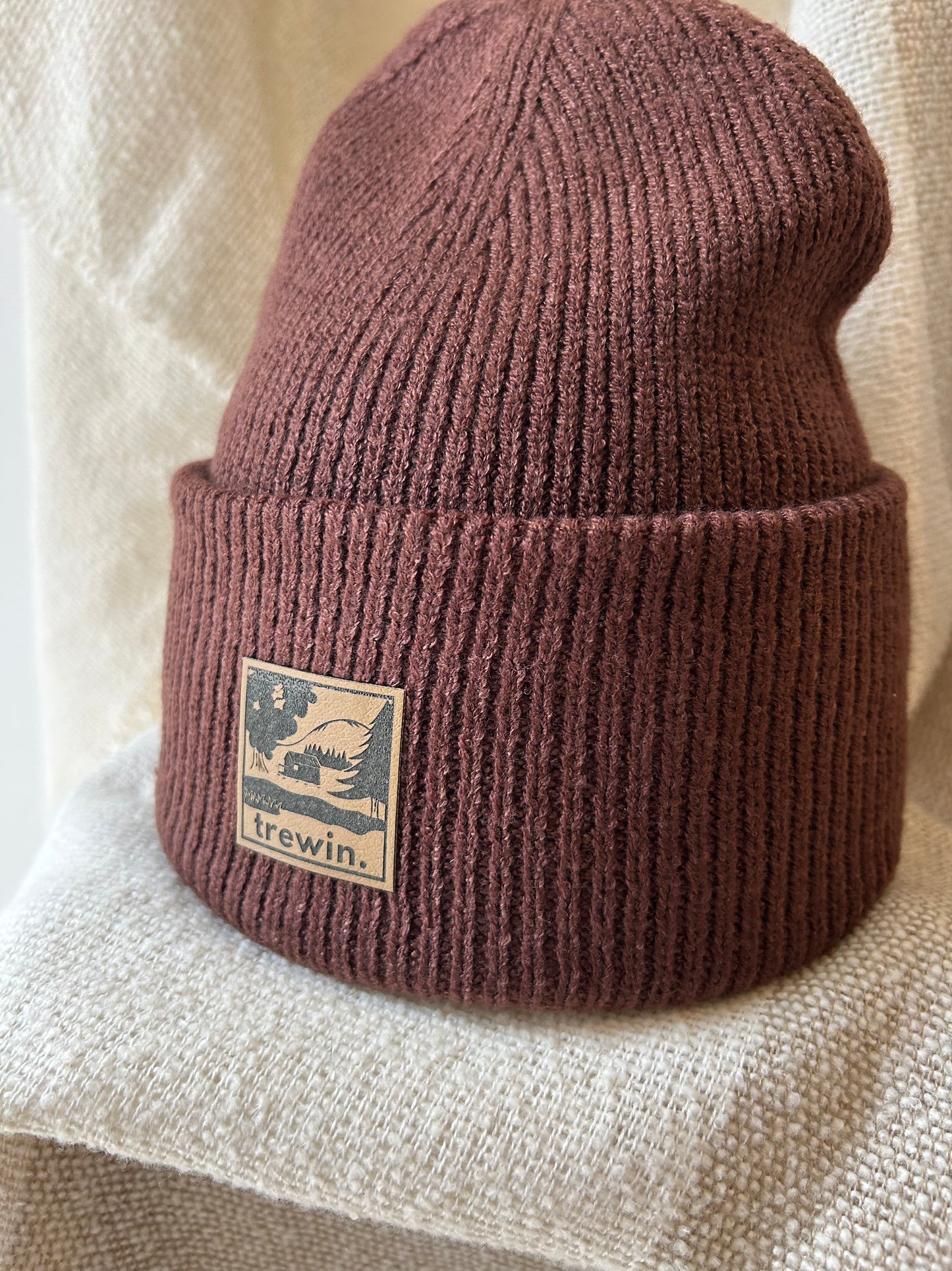 A ribbed knit cuffed beanie in a muted dusty rose or mauve-brown color, featuring a square tan brand patch on the cuff that reads 'irewin.' with a bird logo, displayed on a textured cream fabric background.