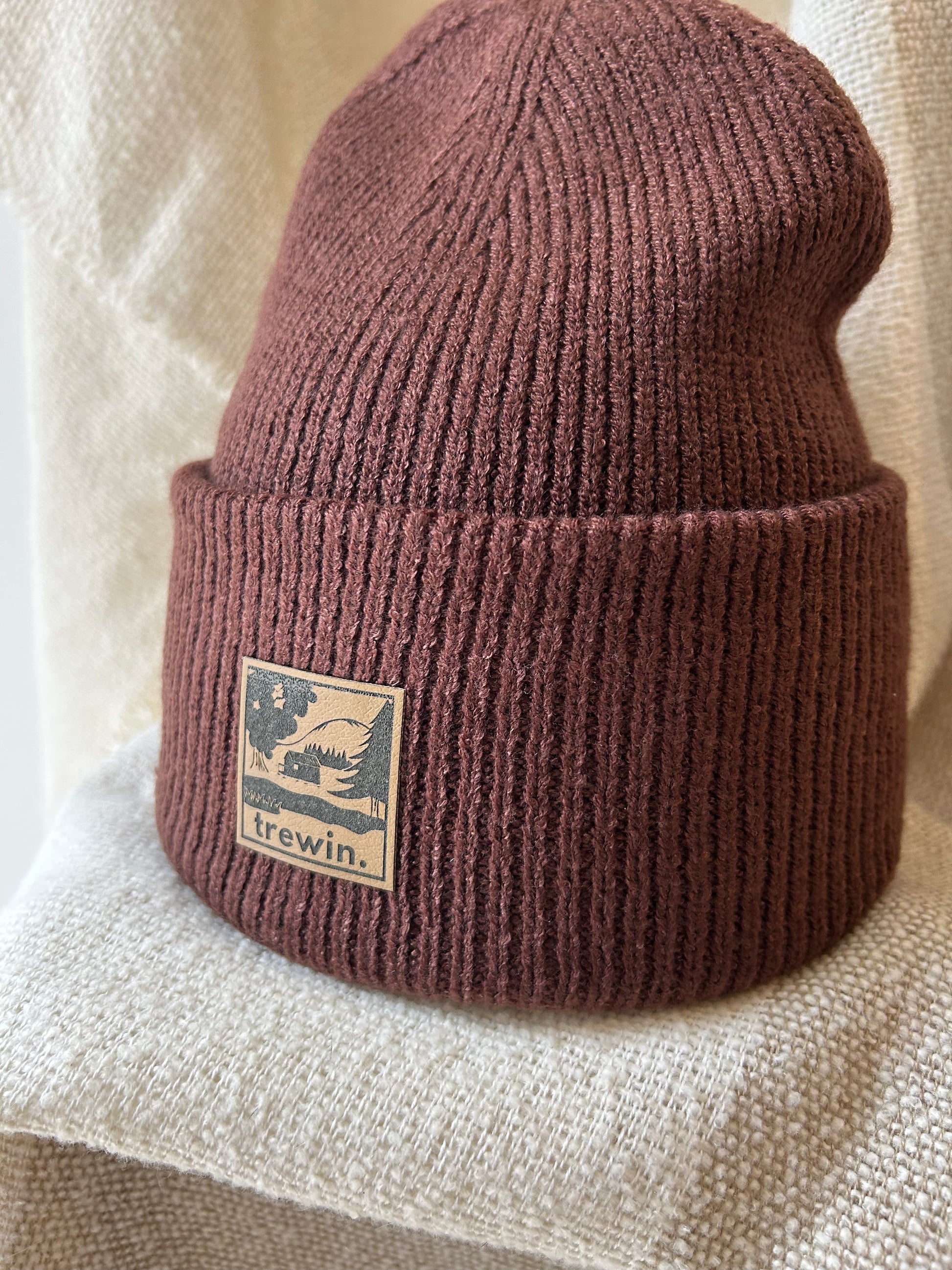 A ribbed knit cuffed beanie in a muted dusty rose or mauve-brown color, featuring a square tan brand patch on the cuff that reads 'irewin.' with a bird logo, displayed on a textured cream fabric background.