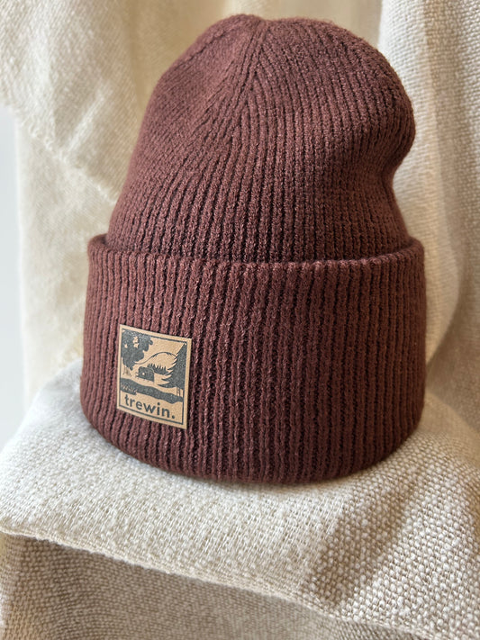 A ribbed knit beanie in a muted dusty rose or mauve-brown color, featuring a square tan brand patch on the front cuff that reads 'trewin.' with a mountain and tree graphic.
