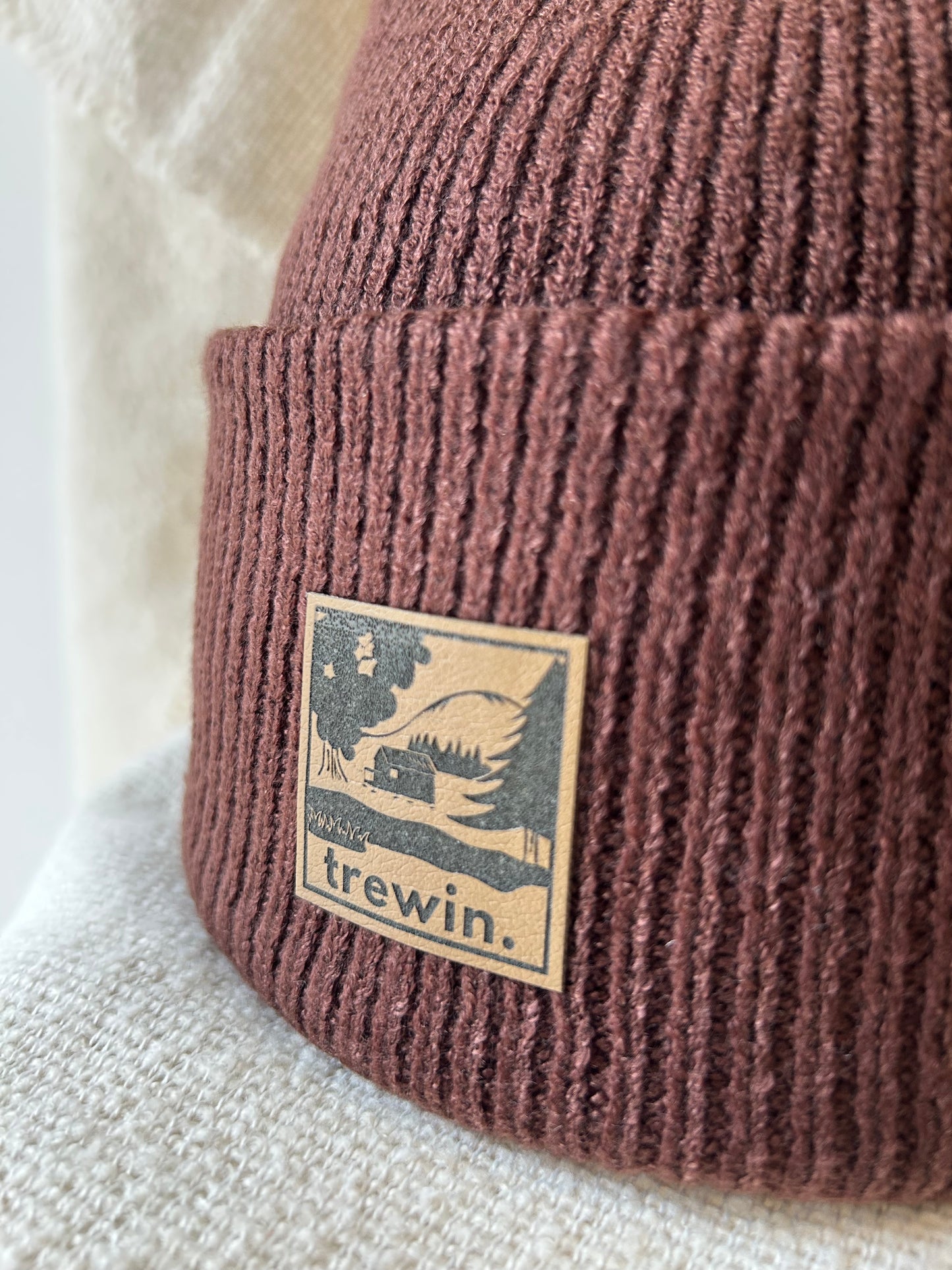 A close-up of a ribbed knit beanie in a dusty mauve-brown color, featuring a square tan patch with a black graphic of a cabin and trees and the text 'trewin.'.
