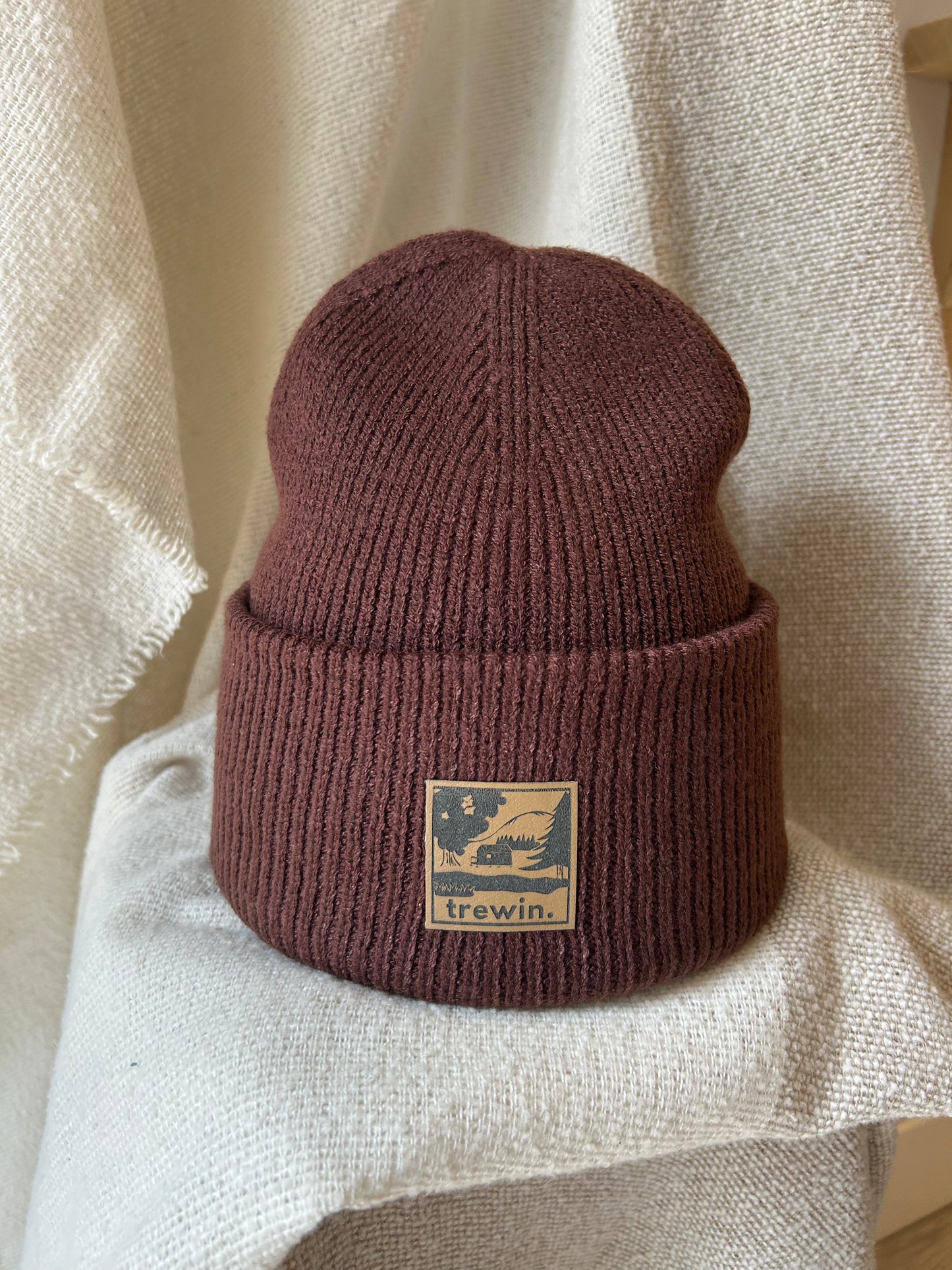 A ribbed knit beanie hat in a muted dusty rose or mauve-brown color, featuring a tan square patch on the front cuff with the brand name 'trewin.' and a graphic of a bird in a tree.