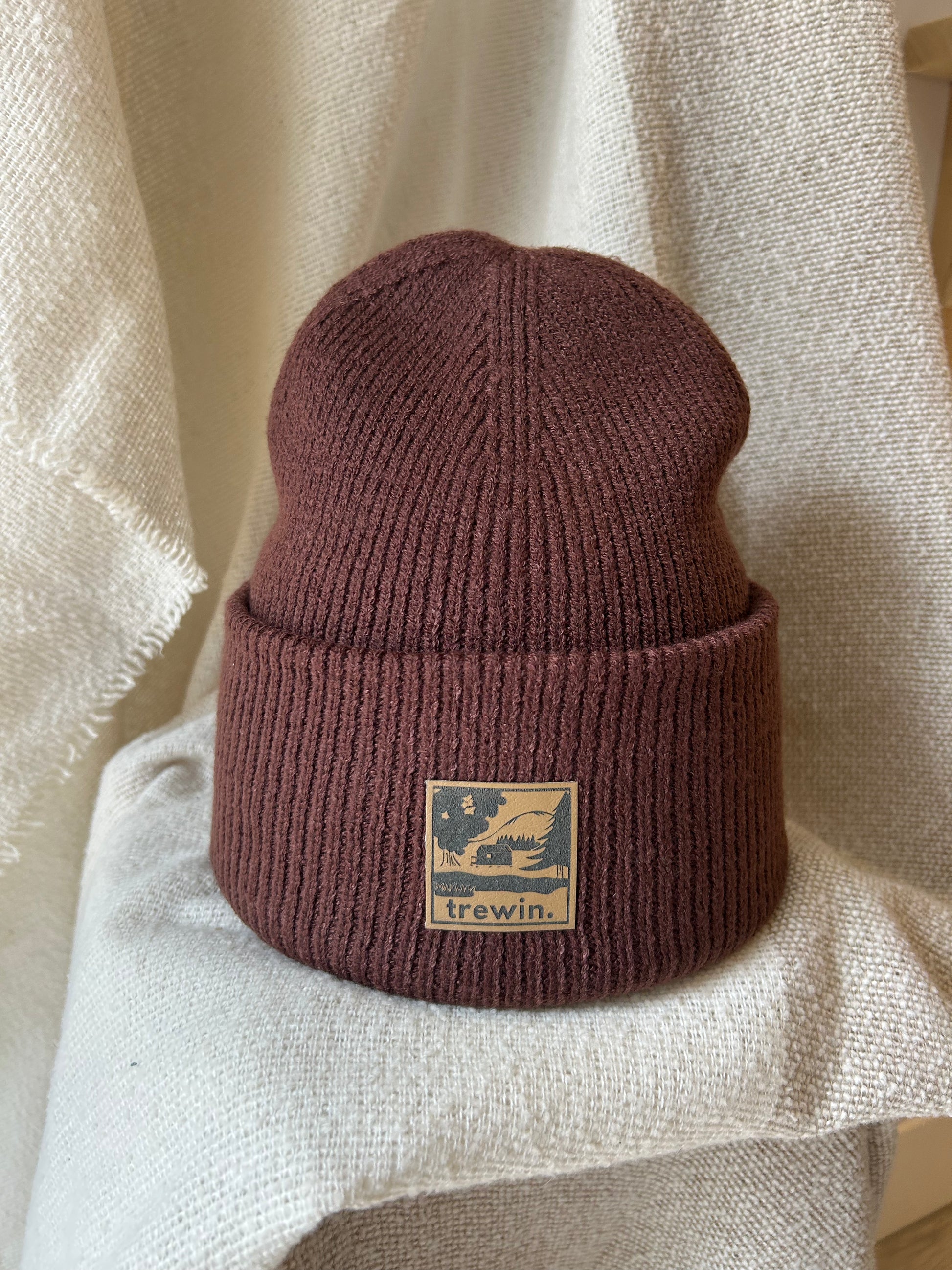 A ribbed knit beanie hat in a muted dusty rose or mauve-brown color, featuring a tan square patch on the front cuff with the brand name 'trewin.' and a graphic of a bird in a tree.