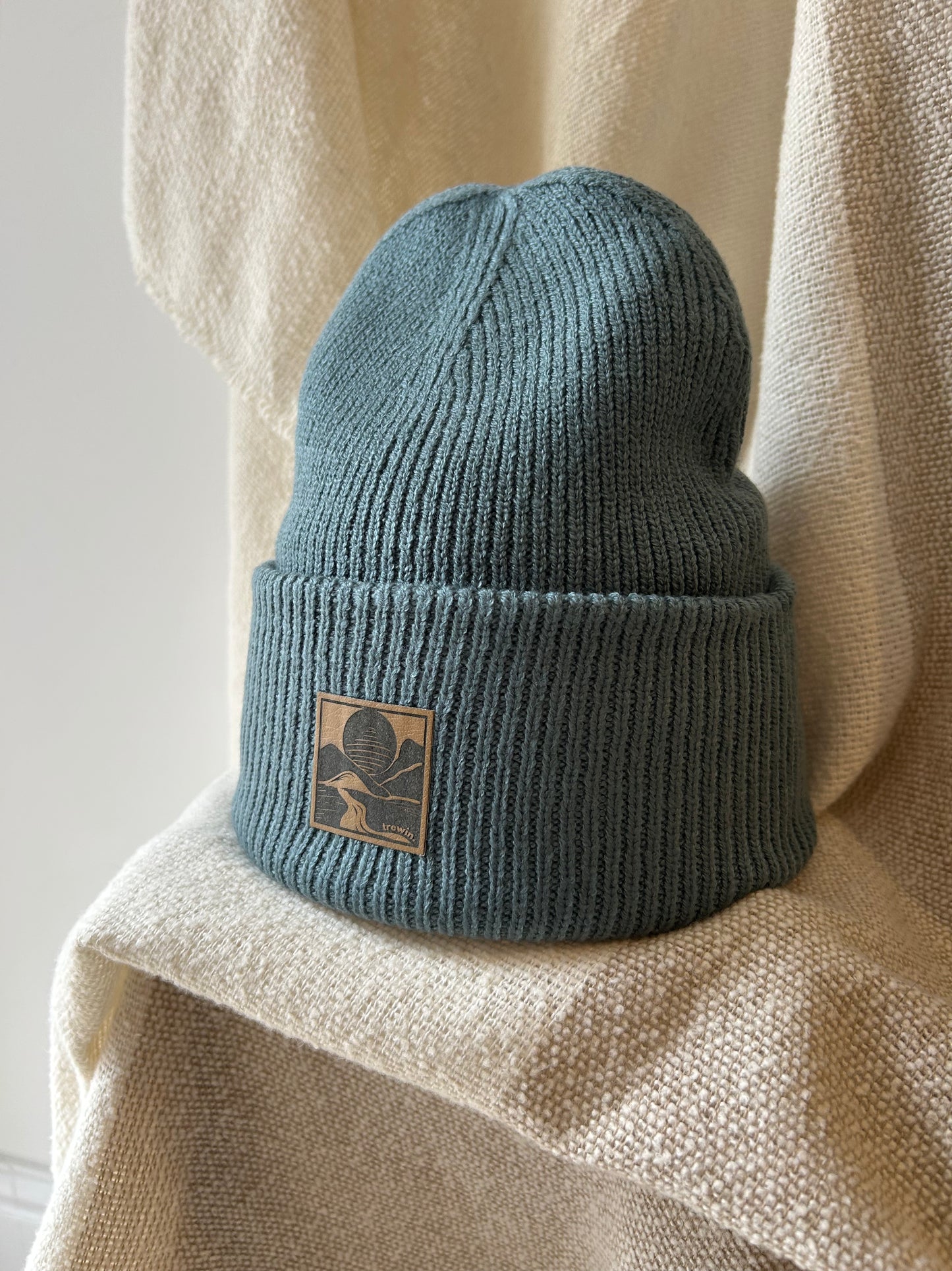 A ribbed knit beanie in muted blue-green, folded over at the brim with a square tan patch featuring a stylized tree logo. The hat is styled on draped cream fabric.