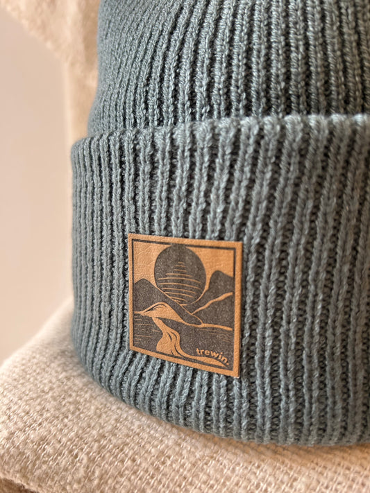 Close-up of a ribbed knit beanie in a muted blue-gray color, featuring a square tan leather patch with a mountain and sun design and the brand name 'browin'.