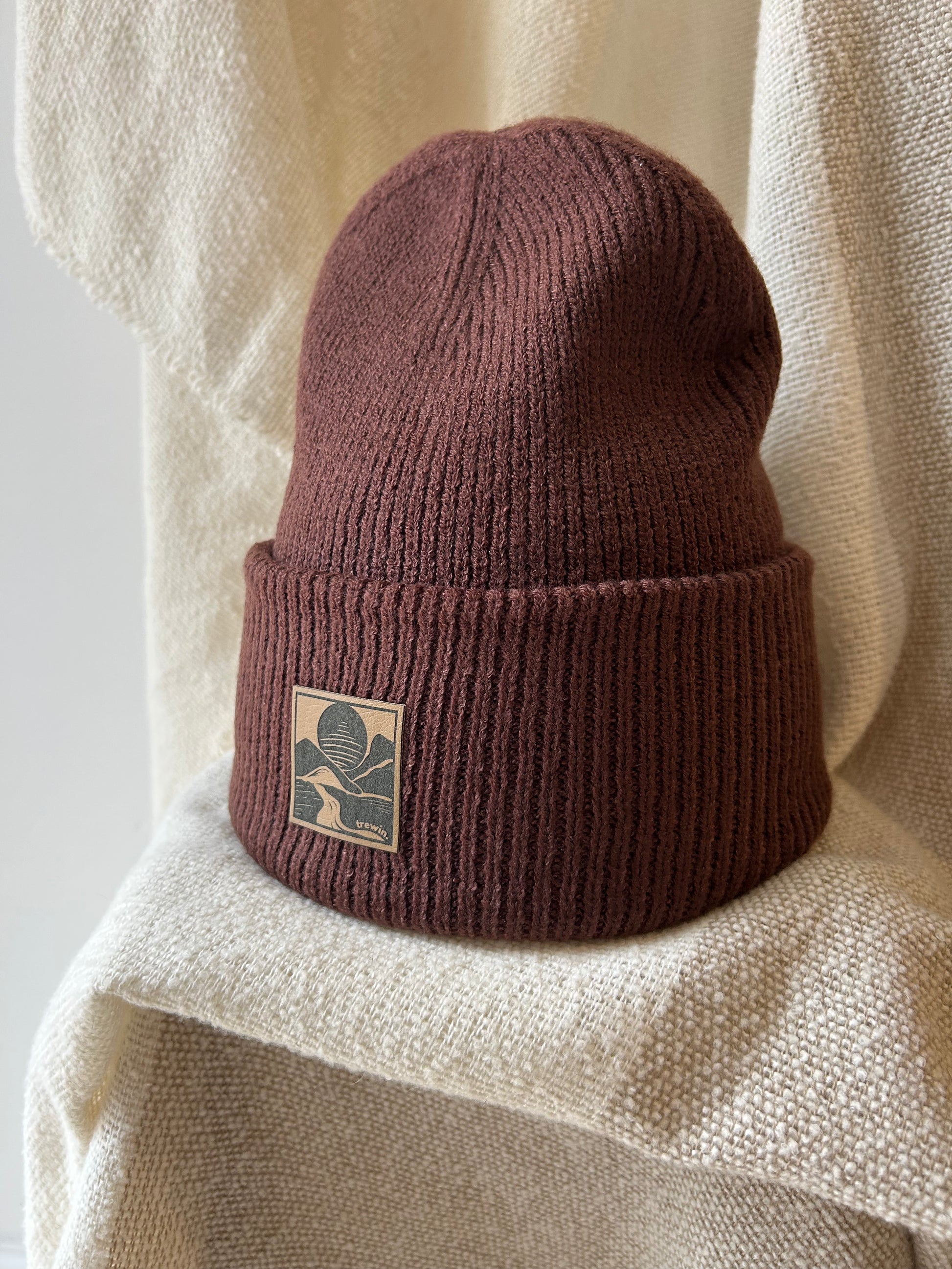 A ribbed knit beanie in a muted burgundy-brown color, featuring a square tan patch on the folded cuff with a dark leaf logo. The hat is styled on draped cream-colored textured fabric.