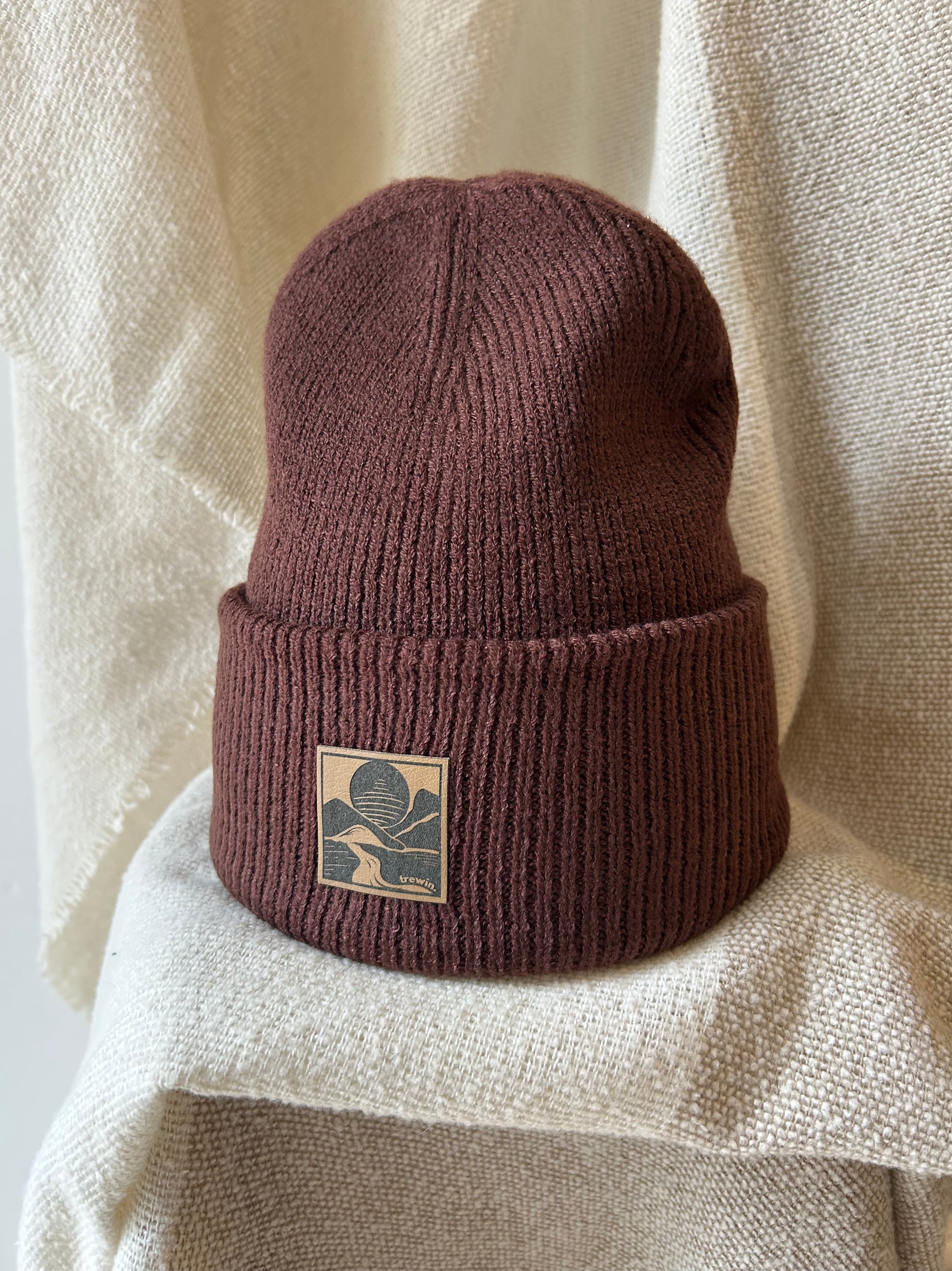 A ribbed knit cuffed beanie in a muted burgundy-brown color, featuring a square tan patch with a dark leaf logo on the front, displayed against a draped beige fabric background.