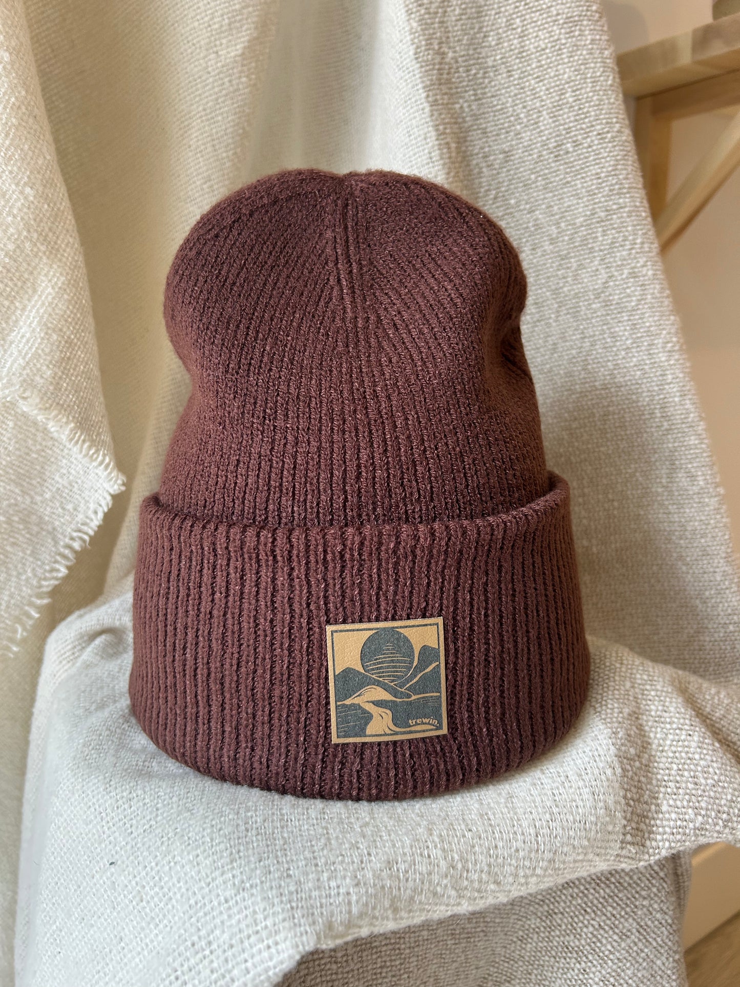 A ribbed knit beanie in a muted dusty rose or mauve-brown color, featuring a square tan patch with a dark blue graphic on the front, displayed on draped cream fabric.