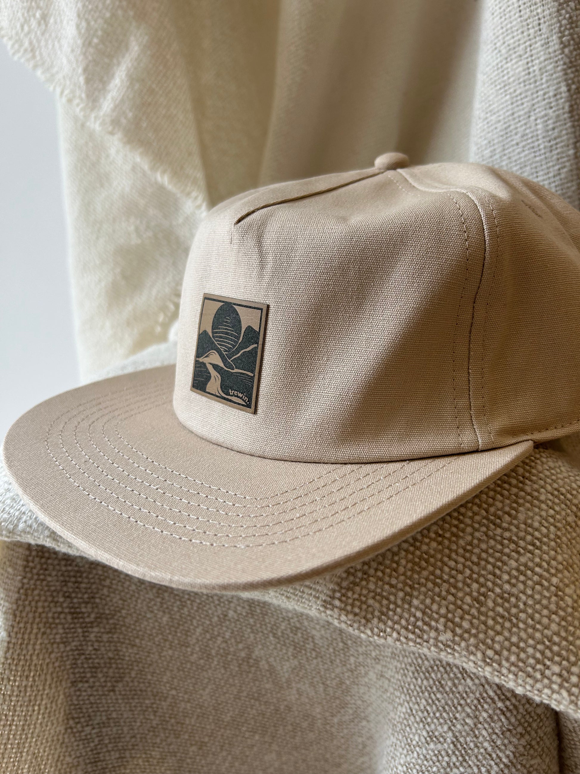 A beige flat-brim cap with a square embroidered patch on the front, styled on a textured cream fabric background.