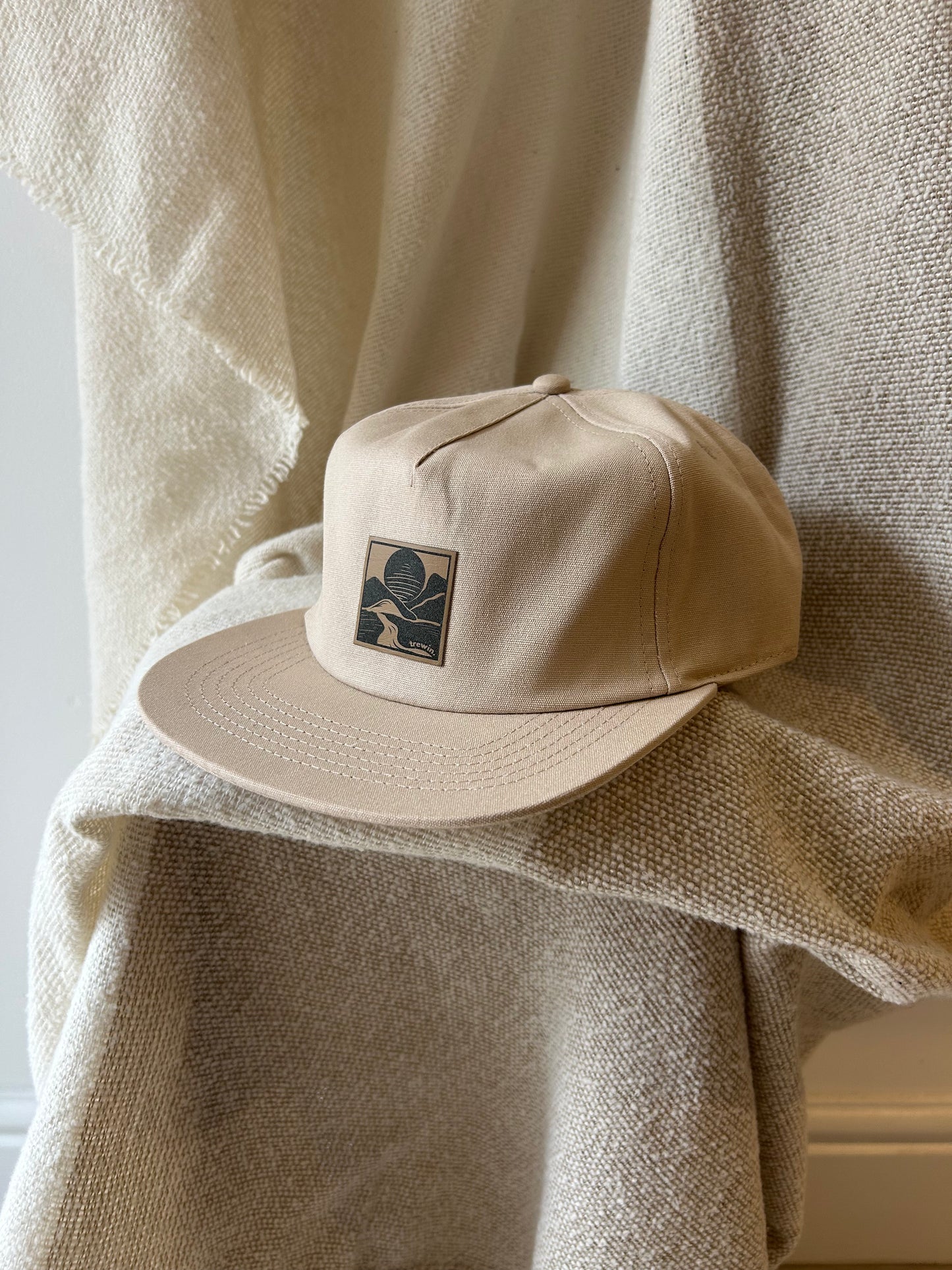 A beige baseball cap with a small embroidered square logo patch, resting on draped cream-colored textured fabric.