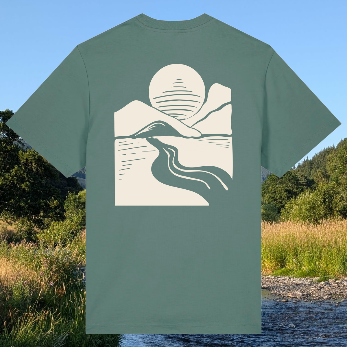 A sage green short-sleeve t-shirt with a cream-colored minimalist graphic of a mountain, sun, and winding river on the back, shown against a background of a natural outdoor landscape with a stream and trees.